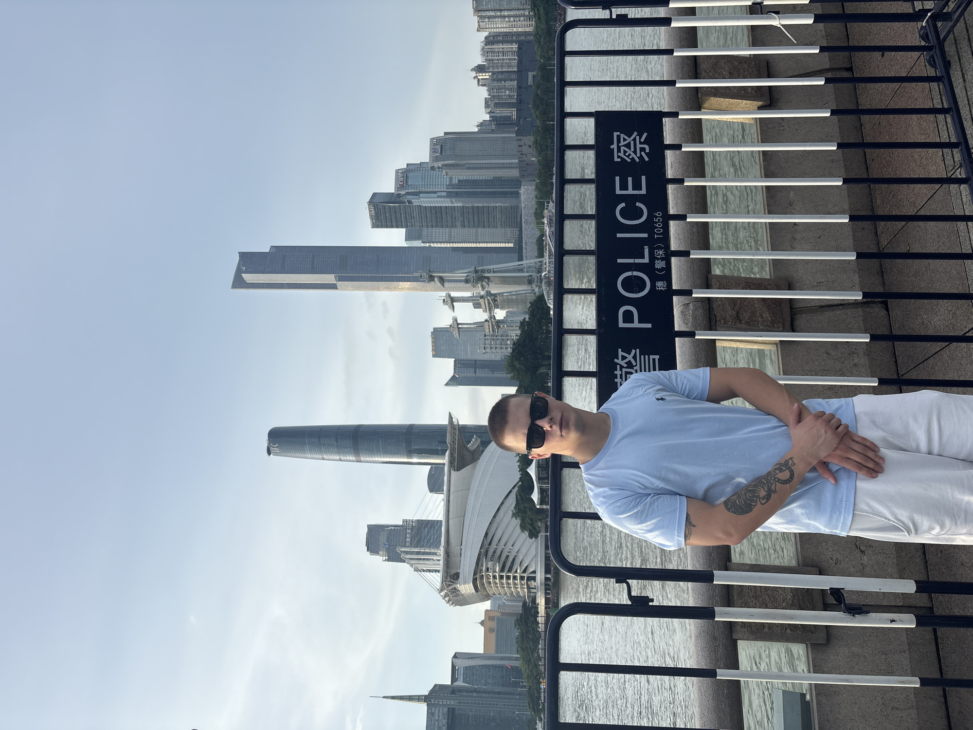 A young Caucasian man stands in front of a metal barrier, with a cityscape of tall buildings and a body of water in the background. The man is wearing sunglasses, a light blue t-shirt with a small logo, and white shorts. He has a tattoo on his left arm. Behind him, a black sign with white lettering reads "POLICE" in English and Chinese characters. The cityscape includes modern skyscrapers, some with distinctive architectural designs, and a large stadium-like structure. The sky is clear with some light clouds. The scene appears to be a waterfront promenade in the Haizhu District of China, likely Guangzhou, given the prominent landmarks.