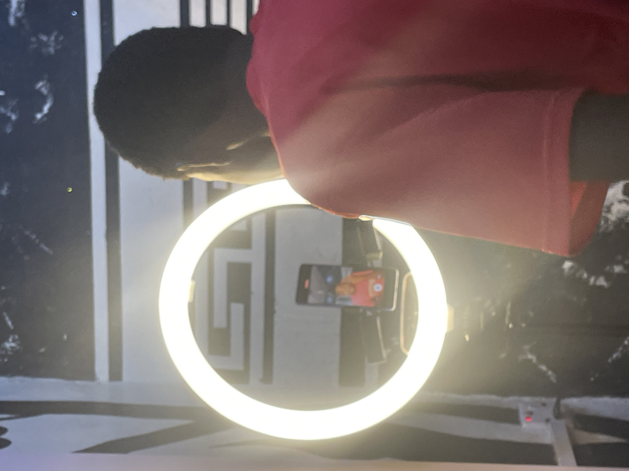 This image captures a close-up, tilted view of a person interacting with a video recording setup in an indoor environment, likely in Onitsha, Nigeria. The main subject is partially visible, showing the back of a person's head with dark hair and their right shoulder, clad in a reddish-pink short-sleeved top. They are positioned in front of a large, circular LED ring light that emits a bright, warm glow, serving as the primary light source.
Centered within the ring light is a horizontally mounted smartphone. Its screen is active, displaying a live camera view or a video recording interface, with a person's face and upper body faintly visible on the display. The background features a wall decorated with a striking black and white geometric pattern, incorporating vertical lines and abstract shapes. In the lower right corner, a white electrical outlet with a small red indicator light is visible, suggesting the ring light is powered on. The scene strongly implies an activity related to video content creation, such as vlogging, live streaming, or a video call. The artificial lighting and equipment suggest this activity could be taking place at any time of day. No legible text is visible in the frame.