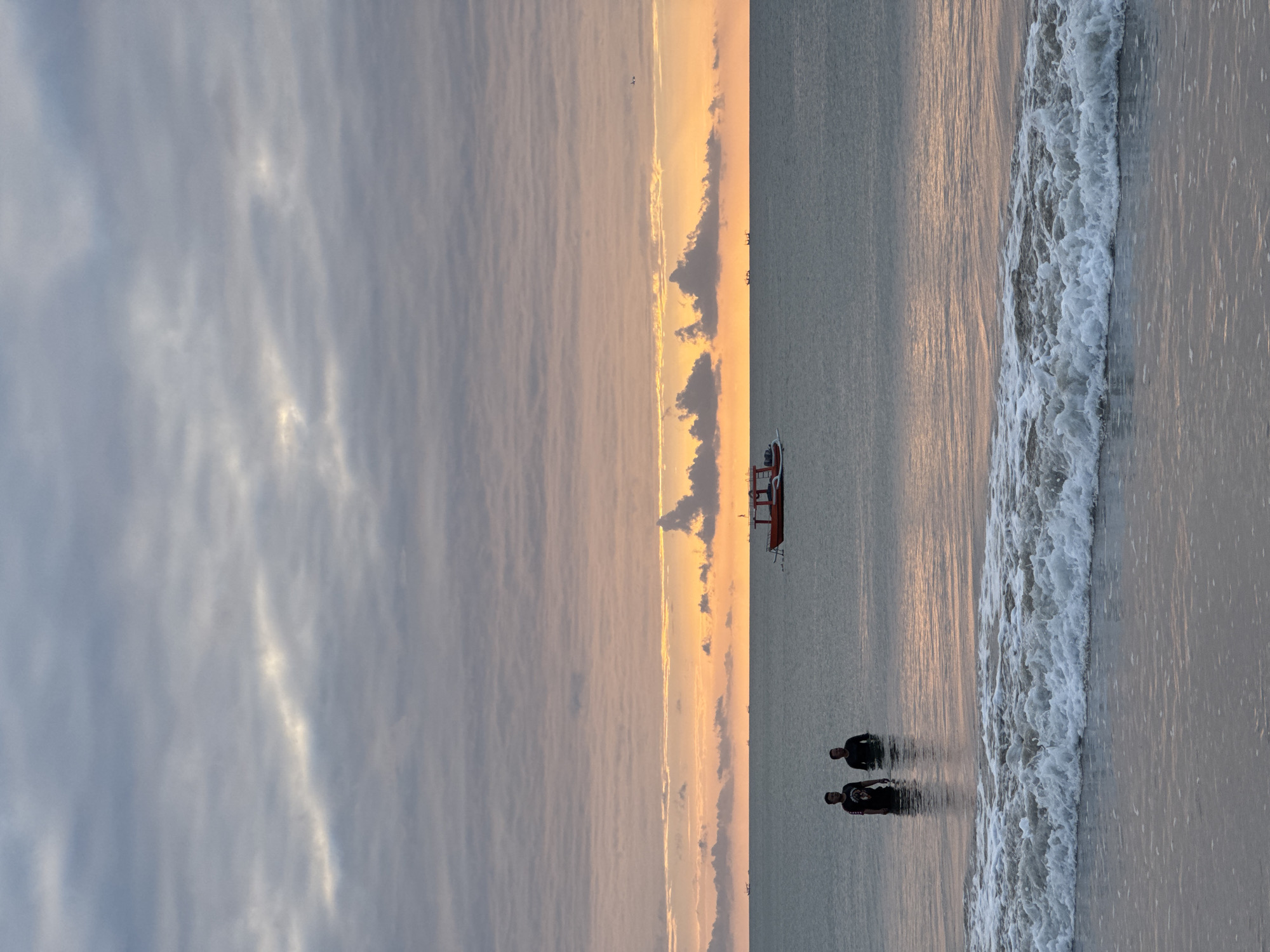 The image depicts a serene seascape at dusk, likely in Kuta Selatan, Indonesia. Two individuals stand in shallow water near the shore, appearing to be interacting or simply enjoying the view. In the distance, a small red boat is anchored, and several other vessels are faintly visible on the horizon. The sky is a canvas of soft orange and grey hues, with textured clouds indicating the setting sun. The ocean is calm, with gentle waves lapping at the shore, reflecting the warm tones of the sky. The overall atmosphere is peaceful and tranquil, capturing a moment of quiet contemplation by the sea.