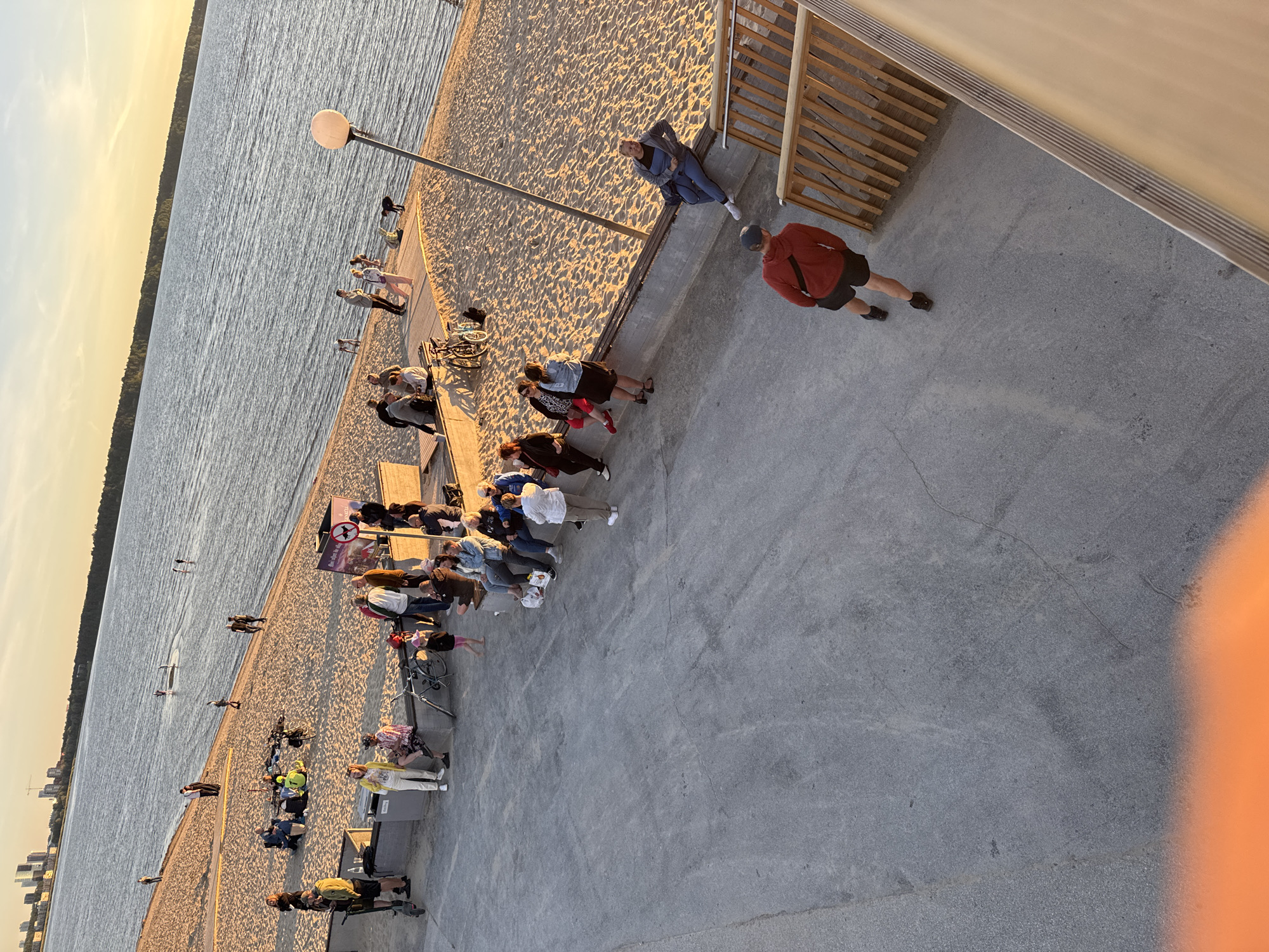 The image depicts a scene at a beach area in Tallinn, Estonia, likely during the late afternoon or early evening, given the warm, golden light casting long shadows. The foreground is dominated by a gray concrete pathway, with a slight orange glow visible at the very bottom edge, possibly from a reflection or artificial light.
Further into the image, a sandy beach stretches along a large body of water, which appears to be a bay or lake. The water is calm, with a few people visible on paddleboards further out. Along the edge of the beach, a wooden walkway or seating area is present.
Numerous people are scattered throughout the scene. On the beach, some are sitting in groups, others are standing, and a few are walking. There are bicycles and personal scooters visible. Near the water's edge, several individuals are standing or sitting. On the concrete pathway, a man in a red hoodie and black shorts is walking away from the camera. On a raised wooden structure with stairs, a woman in a blue jumpsuit is seated.
In the background, across the water, a city skyline with modern buildings is visible, suggesting the beach is in close proximity to an urban area. The sky transitions from a bright yellow-orange near the horizon to a lighter blue above, indicating the setting sun. The overall atmosphere appears relaxed, with people enjoying the outdoor space and the evening light.