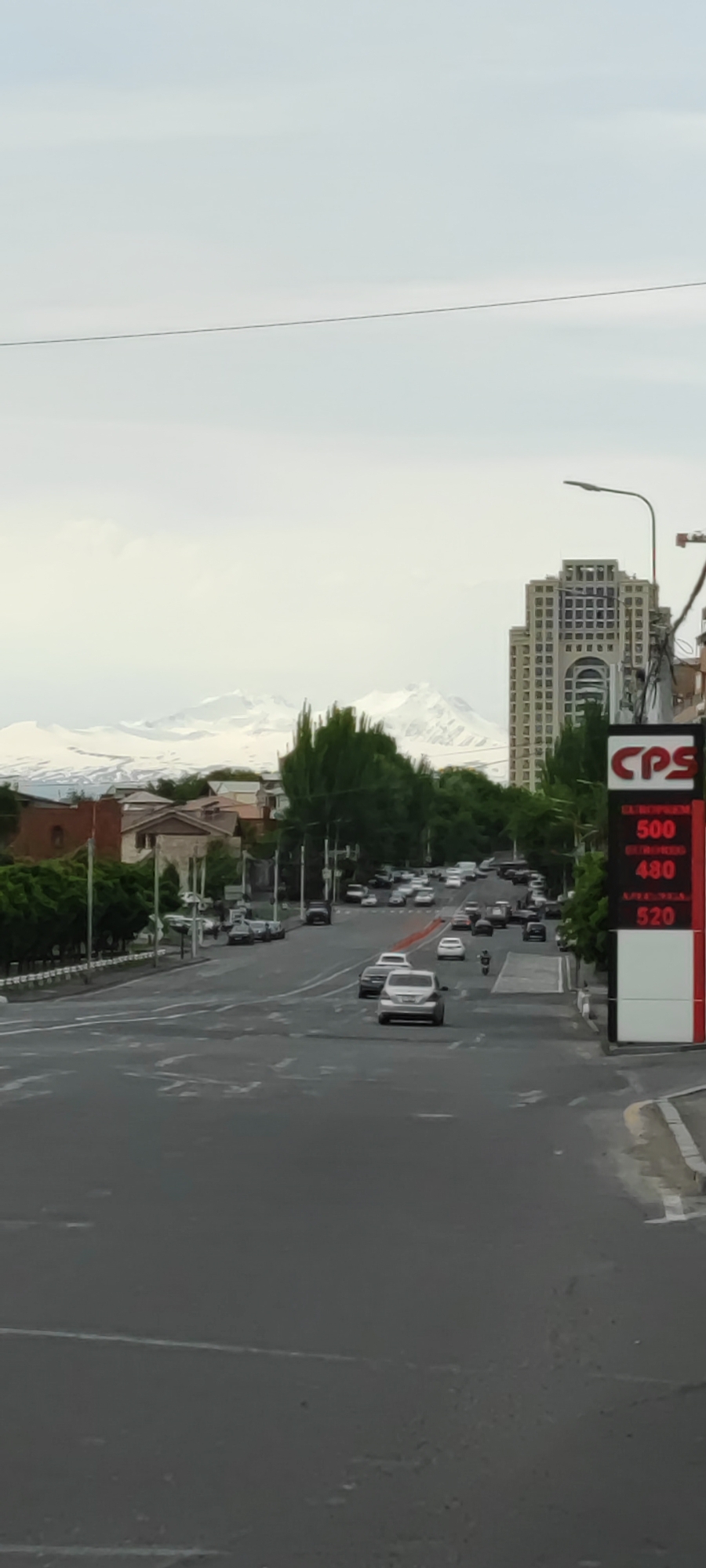 This is a street scene in Yerevan, Armenia. The image is shot from a slightly elevated position looking down a multi-lane road with moderate traffic. The road is asphalt, marked with white lane dividers, and has some minor wear and tear. In the background, a snow-capped mountain range is visible. To the right, there is a tall, modern building with architectural details. Closer to the right is a large, red and white sign with the letters "CPS" and some numbers, indicating it could be a gas station sign showing fuel prices. Trees line the sides of the street, and there are other buildings visible. The sky is overcast, and the overall lighting is somewhat flat. In the middle ground, there are several cars and one motorcycle moving along the road. No pedestrians are clearly visible in the immediate foreground.