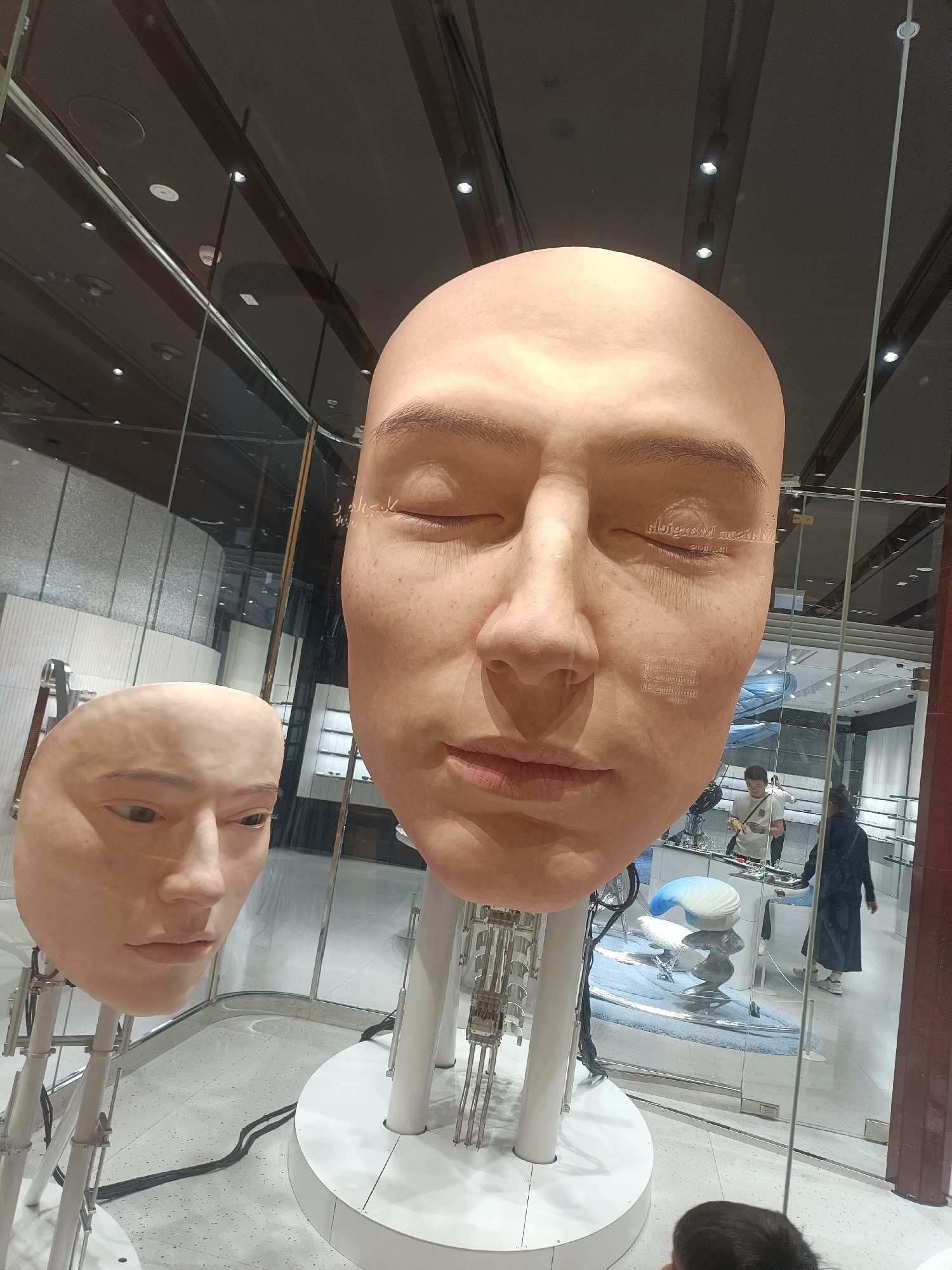 The image shows two large, realistic human heads as part of an art installation or display, likely in a retail environment or museum. The head in the foreground is significantly larger and appears to be mounted on a stand with visible mechanical components. This head has closed eyes and subtle freckles on its skin. To the left, a smaller, similarly realistic head is also visible, mounted on a stand. Both heads are made of a pale, skin-toned material. Behind the heads, a reflection shows a store interior with shelves stocked with items, possibly eyewear or accessories, and several individuals browsing or working. The lighting suggests an indoor setting, possibly a well-lit shopping mall or exhibition space. The presence of what appears to be Arabic script on the larger head and the overall aesthetic suggest a location in Dubai, United Arab Emirates, possibly within a luxury retail establishment or a contemporary art exhibition space.