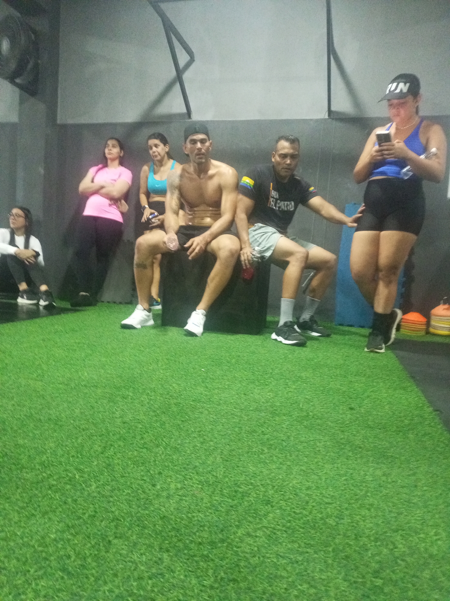 The image depicts a group of individuals in a gym setting, with an artificial turf floor. In the foreground, a man with a bare chest and black shorts sits on a large black object, while another man in a black t-shirt and grey shorts sits next to him. To the left, two women are seated on the floor. One woman, wearing a pink t-shirt and black leggings, has her arms crossed. The other woman, in a white long-sleeved shirt and black leggings, is looking to the side. In the background, a woman in a blue sports bra and black shorts stands looking at her phone, wearing a black cap. Another woman in a pink t-shirt is also visible in the background. The environment suggests a training or break area within the gym, with gym equipment possibly out of frame. The scene appears to be located in the city of Juan Griego, Venezuela.