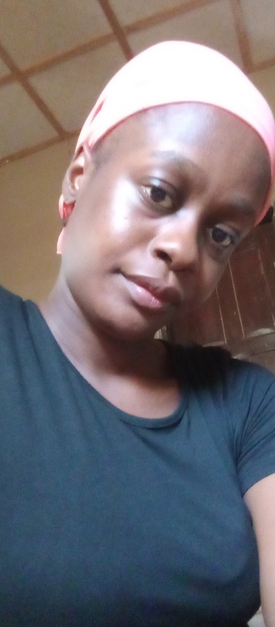 A close-up shot shows a woman with dark skin wearing a peach-colored head wrap and a dark-colored shirt. The background consists of a ceiling with a grid pattern, suggesting an indoor environment. Part of a wooden doorframe is visible on the right side of the frame. The image appears to be taken in a residential setting. The specific location in Akwanga, Nigeria is indeterminable from the image alone.