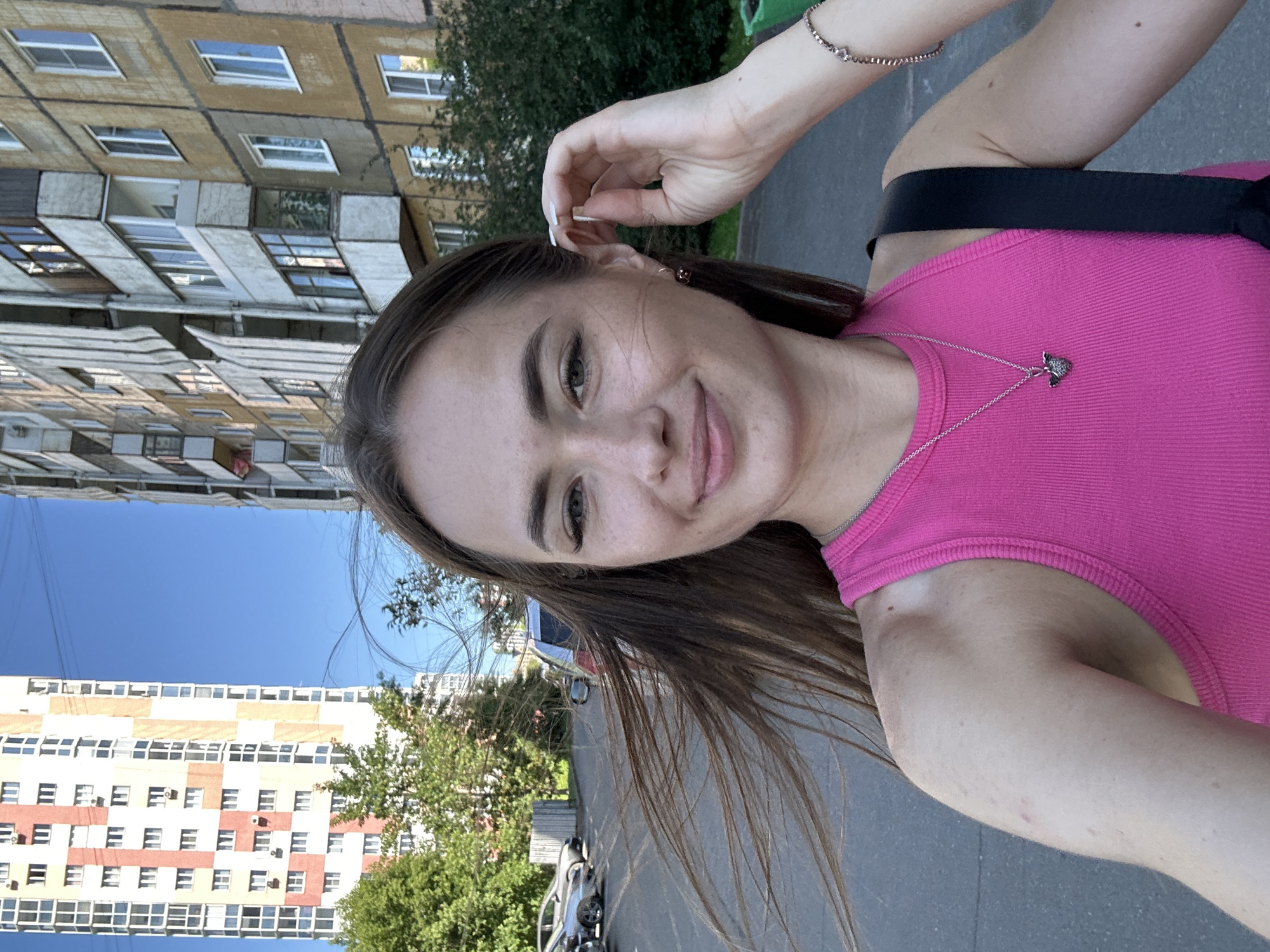 In an outdoor urban setting, a young woman with long brown hair pulled back from her face is taking a selfie. She is wearing a bright pink ribbed tank top and a silver necklace with a dark pendant. Her right hand is raised to her ear. In the background, a tall, colorful apartment building is visible to the left, and a weathered, multi-story apartment building stands to the right. Trees and parked cars are also present. The sky is clear and blue.