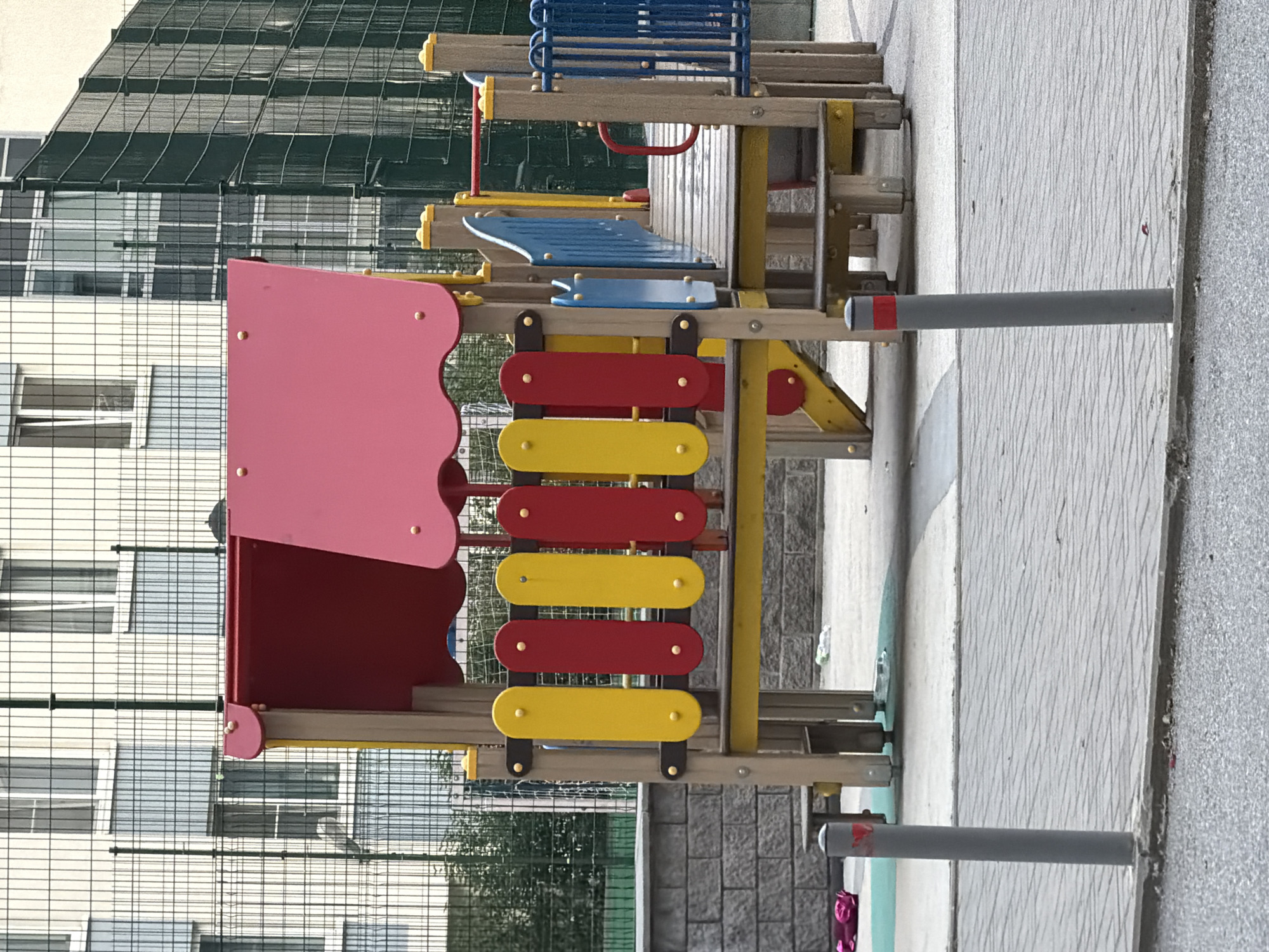 This image captures an empty playground structure within an urban residential area of Saint Petersburg, Russia. The central subject is a brightly colored playground, constructed from wood and plastic, featuring a red and pink multi-level roof. Below the roof, a barrier is formed by alternating vertical yellow and red panels, secured with small yellow rivets. The structure includes wooden support beams and platforms, a wooden bridge section with blue paneled railings on one side, and blue vertical bar railings on the other. A small red handrail is visible on the bridge.
In the background, behind the playground, a tall, dark-green metal mesh fence separates the play area from several multi-story residential buildings. These buildings have light-colored facades and numerous windows, some of which are slightly open. The ground within the playground area consists of light-colored concrete paving, with sections of a soft, light blue/green surface. To the left, a grey stone wall constructed from rectangular blocks is visible.
The foreground features a light-grey paved sidewalk made of interlocking rectangular pavers, separated by a curb from a darker, possibly asphalt, surface. Two grey bollards, each with a red reflective band near the top, are embedded in the sidewalk, marking its edge. Scattered debris includes a crumpled pink item and a plastic bottle on the ground near the playground. No people are present in the scene.