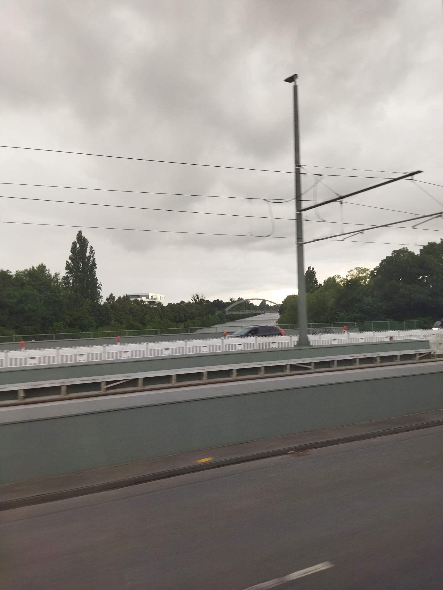 The image captures a view of an elevated structure in Hanover, Germany, likely a highway or railway line, under a cloudy sky. The foreground shows a grey asphalt road. Above the road is a green wall, and behind that, a white barrier with orange markers. Further back, the scene reveals trees and a modern white building in the distance. Power lines and a streetlamp are visible in the sky. Several vehicles, including a dark car and what appears to be a motorcycle, are present on the elevated structure. The overall impression is a contemporary urban landscape during overcast weather.