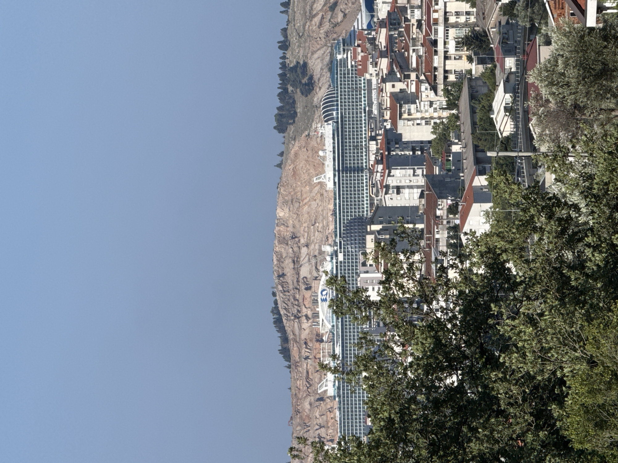This image depicts a scenic view of the city of Sustaš, Montenegro, showcasing a large cruise ship docked in the distance. The cruise ship, prominently white with dark-tinted windows, is positioned behind a cluster of residential buildings. These buildings, characterized by their varied architectural styles and red-tiled roofs, create a textured urban landscape. In the foreground, lush green trees partially obscure the view of the city. Behind the cruise ship and the buildings, a rugged, rocky hillside rises, providing a natural backdrop to the scene. The sky above is a clear, pale blue, suggesting a sunny day.