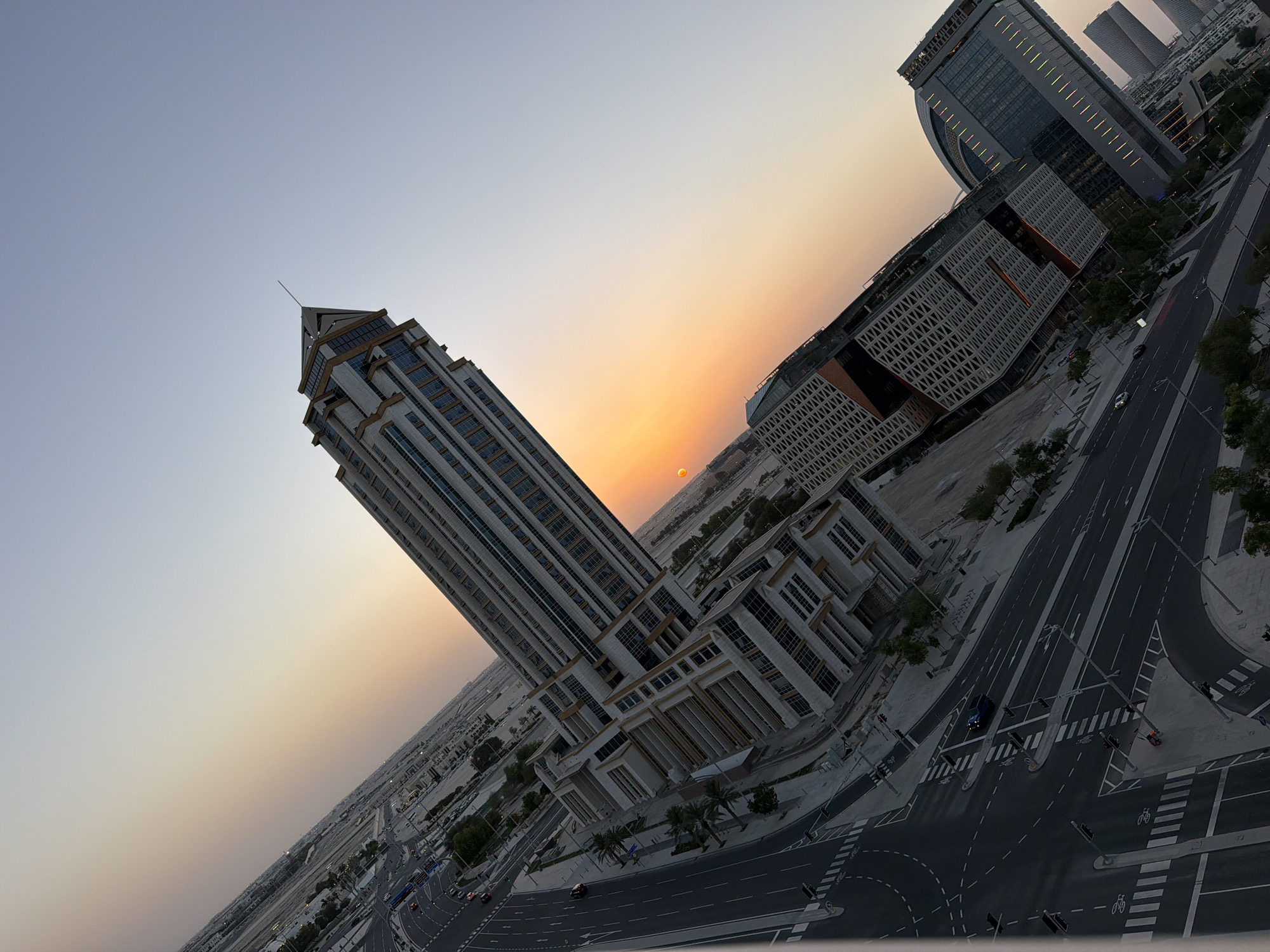This is a tilted, eye-level, outdoor, medium shot photograph of a city at sunset, taken from a high vantage point. The sun, a bright orange orb, is visible on the horizon, casting a warm glow across the sky. In the foreground, a long, multi-story modern building with a sharp, pointed roof dominates the frame, its windows reflecting the sunset hues. To the right of this building, another modern structure, wider and with a curved facade, is partially visible. Streets with traffic, including cars and motorcycles, are depicted below. The environment is urban, with paved roads, sidewalks, streetlights, and some sparse greenery. The sky transitions from a pale blue at the top to a gradient of orange and pink as it nears the horizon.