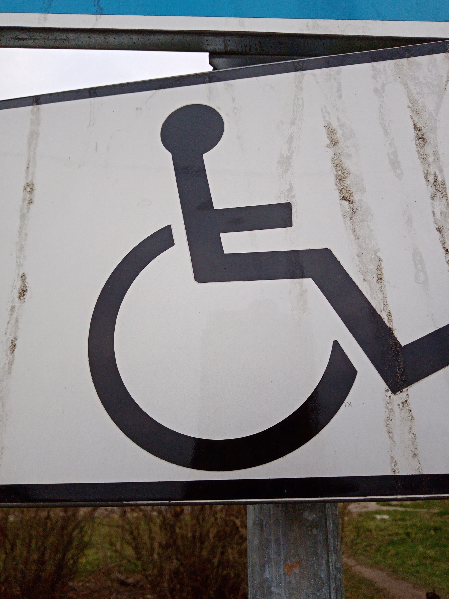 A close-up of a dirty, white sign with a black universal wheelchair symbol. The sign is mounted on a metal pole. The background includes blurry foliage. The sign is located in Elektrostal, Russia.