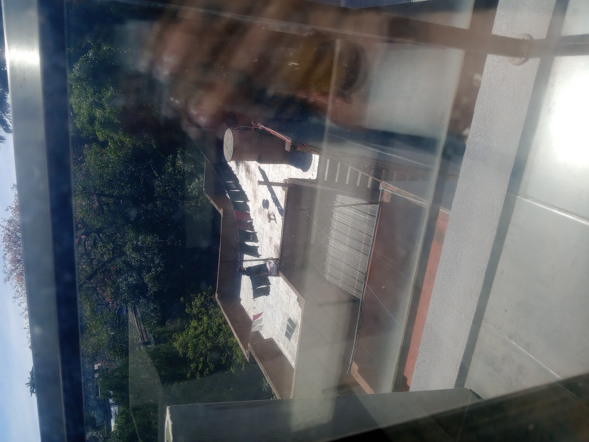 This image, taken from an elevated position such as an upper floor window, shows a building in Lomas de Zamora, Argentina. The view is partially obscured by reflections on the glass.
The building appears to be a multi-story structure with a light-colored facade and a corrugated metal roof. Laundry lines are visible on the roof, with clothes hanging to dry. The building has a cylindrical structure on its roof.
Adjacent to the building is a dense area of trees and foliage. The sky is partially visible in the upper left corner. The immediate foreground consists of the window frame. The angle of the shot suggests a downward-looking perspective.