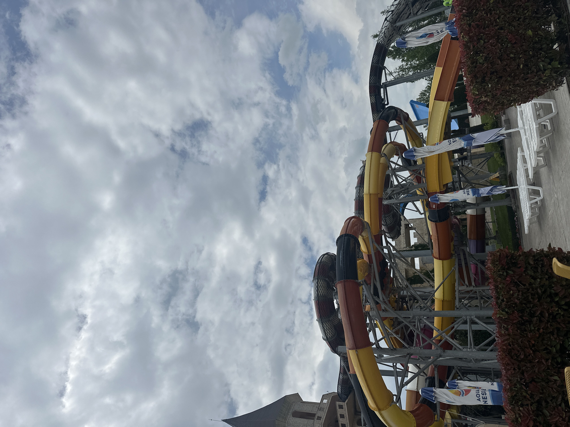 The image depicts a water park attraction against a backdrop of a cloudy sky. The location is Ravda, Bulgaria. There are multiple intertwined water slides, predominantly in yellow, orange, and brown colors. Supporting the slides is a metal framework. Partially visible in the foreground are buildings with pointed roofs. In the background, there are white sun loungers and umbrellas. The composition suggests an outdoor setting, likely a recreational or tourist location. No people are visible in the image.