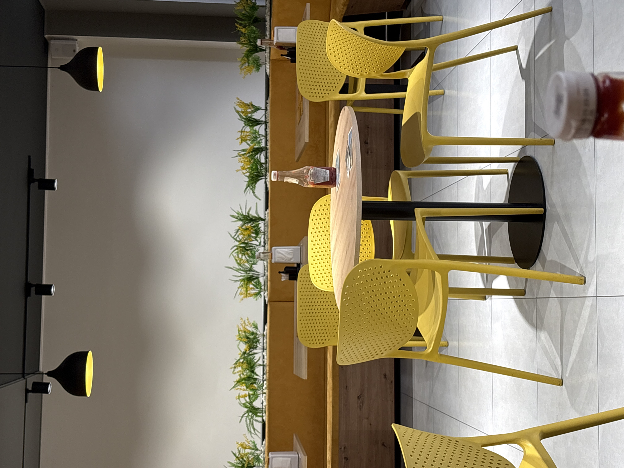 The image shows a restaurant interior with yellow plastic chairs arranged around a light-colored wooden table. A bottle of ketchup sits on the table. Green plants line the wall in the background, and the space is lit by black pendant lights. The scene is located in Yerevan, Armenia.