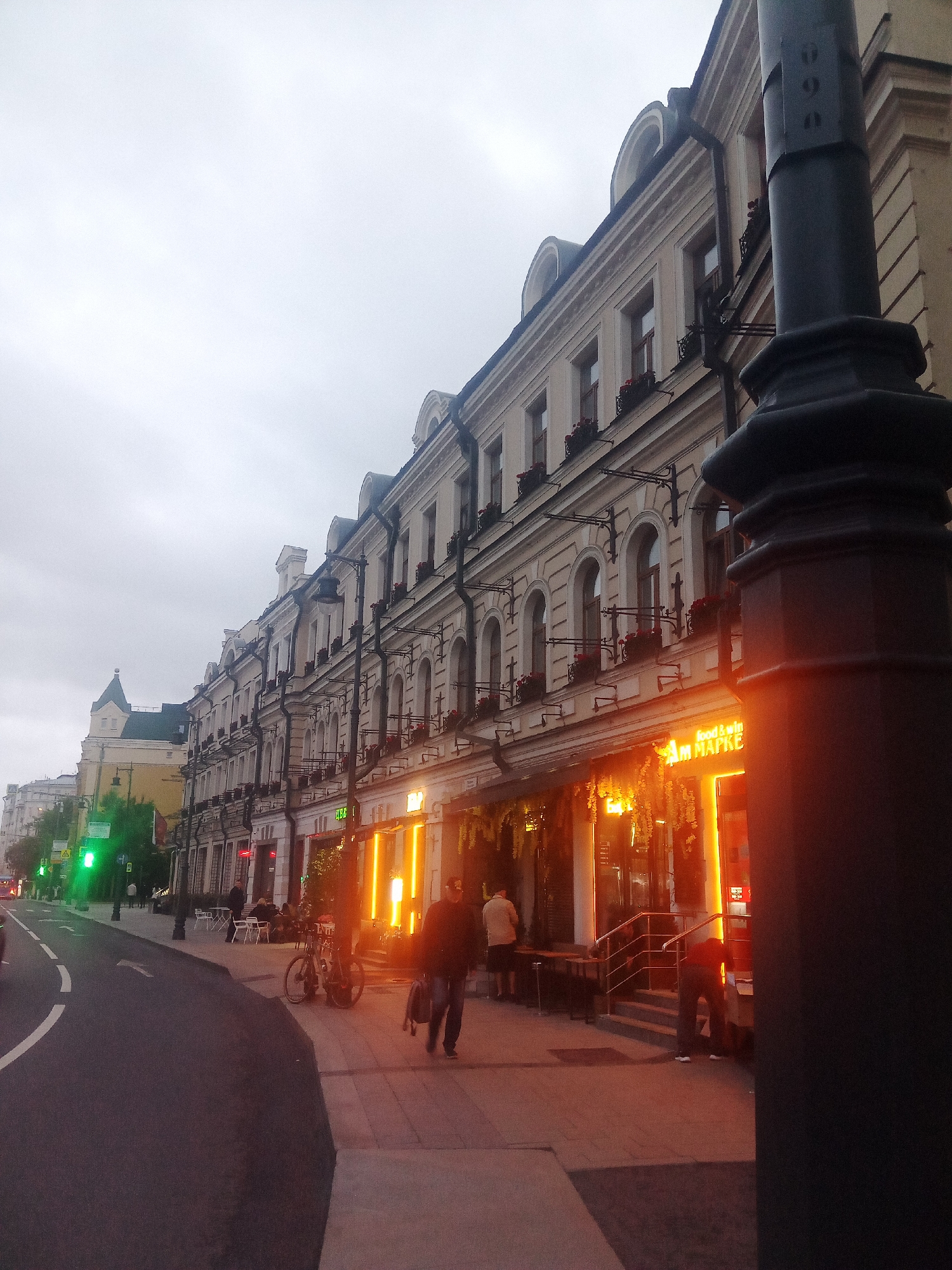 The image depicts a street scene in Moscow, Russia, likely in the evening or overcast weather. A long, ornate building with multiple floors and arched windows lines the right side of the street. The building is painted a light color and has dark window frames and decorative elements. A large, dark lamppost is prominent in the foreground on the right.
On the sidewalk in front of the building, several people are visible. Two individuals are walking towards the viewer, one carrying a bag. Another person appears to be bending down near the entrance of a brightly lit establishment. There are tables and chairs set up outside this establishment, suggesting it is a cafe or restaurant. A bicycle is parked on the sidewalk.
The street itself curves away to the left, with a lane marked by a white line. A green traffic light is visible on the left side of the street. Further down the street, more buildings and traffic can be seen, though they are less distinct due to the distance and lighting conditions. The sky is overcast and grey. The overall impression is of a moderately busy urban street.