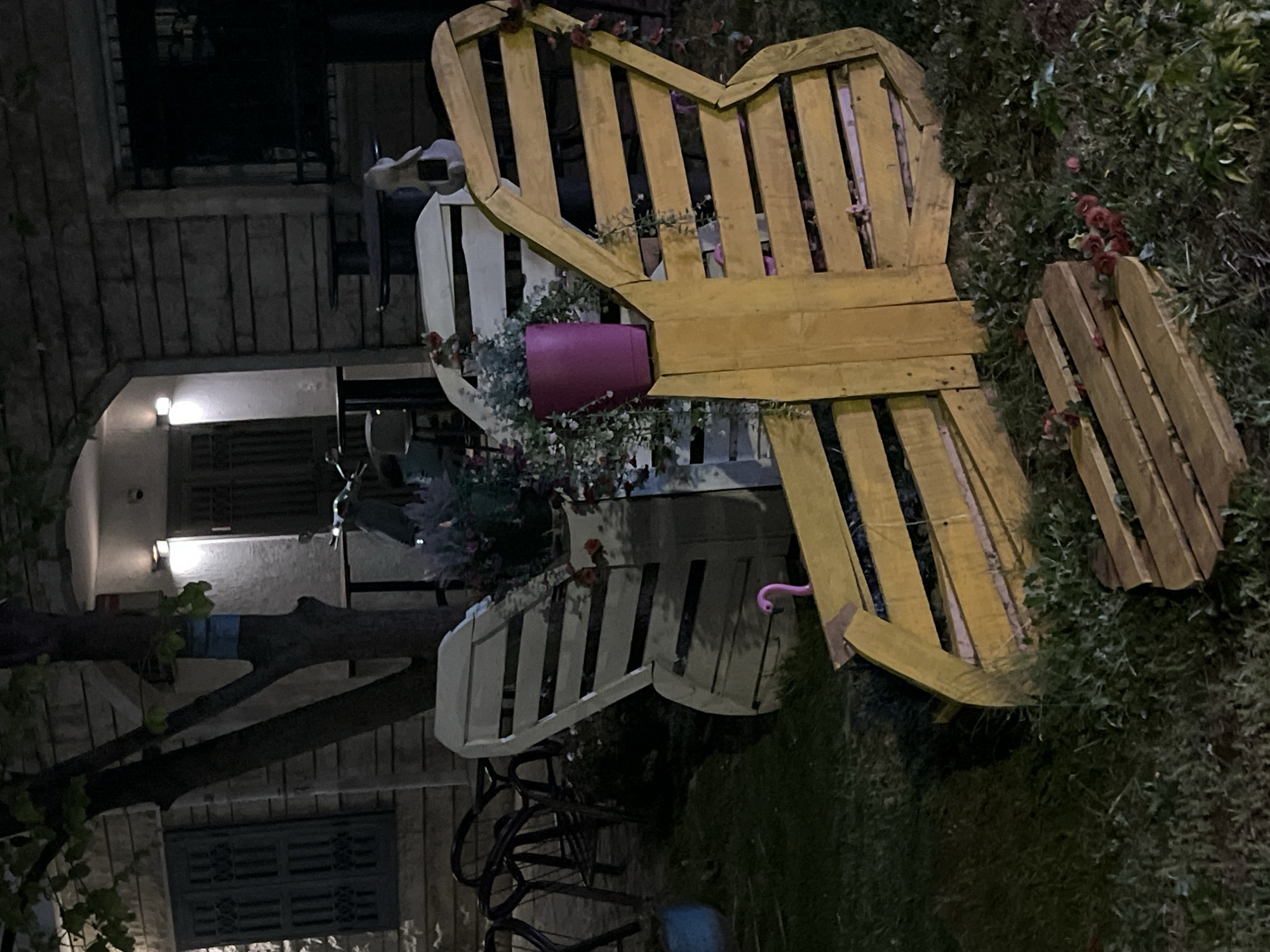 The image depicts a garden scene in Budva, Montenegro. A yellow wooden heart-shaped bench is prominently featured, next to a white wooden bench. Both are adorned with flowers and a pink flower pot. Adjacent to the bench is a portion of a building with a tree extending from it, and windows with closed shutters. The setting appears to be an outdoor area, potentially a private garden or yard. The lighting suggests it is night. Vegetation, including grass and other greenery, is visible around the bench. Another yellow wooden structure, possibly part of a set of furniture, is situated behind the main bench.