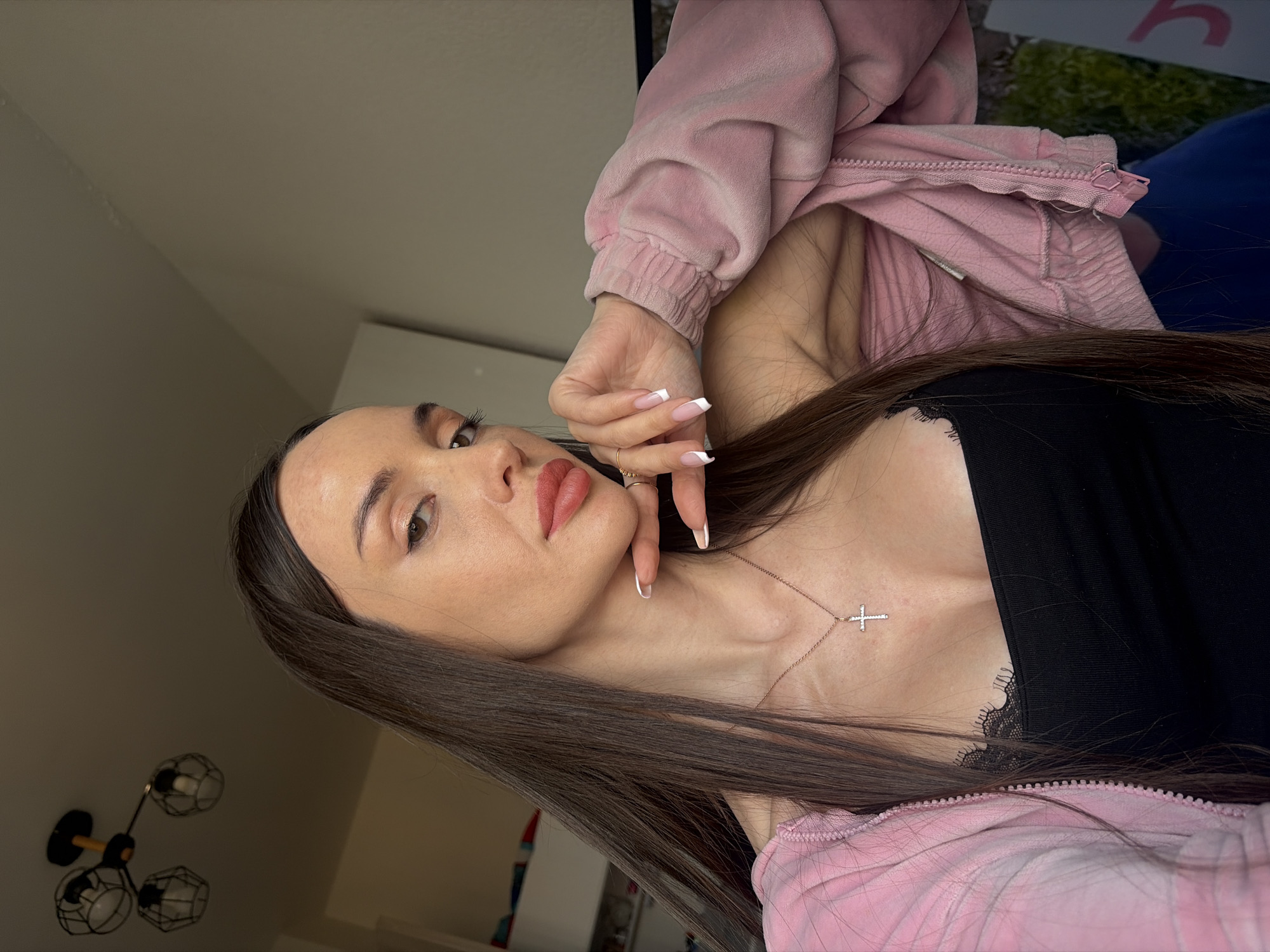 A woman is posing indoors. She has long brown hair and is wearing a black tank top with lace trim and a pink jacket. She is also wearing a gold necklace with a cross pendant. Her fingernails are manicured with a French tip. The background is blurry, but there is a glimpse of a light fixture with a geometric design. There is insufficient information to determine if the image was taken in Saint Petersburg, Russia.