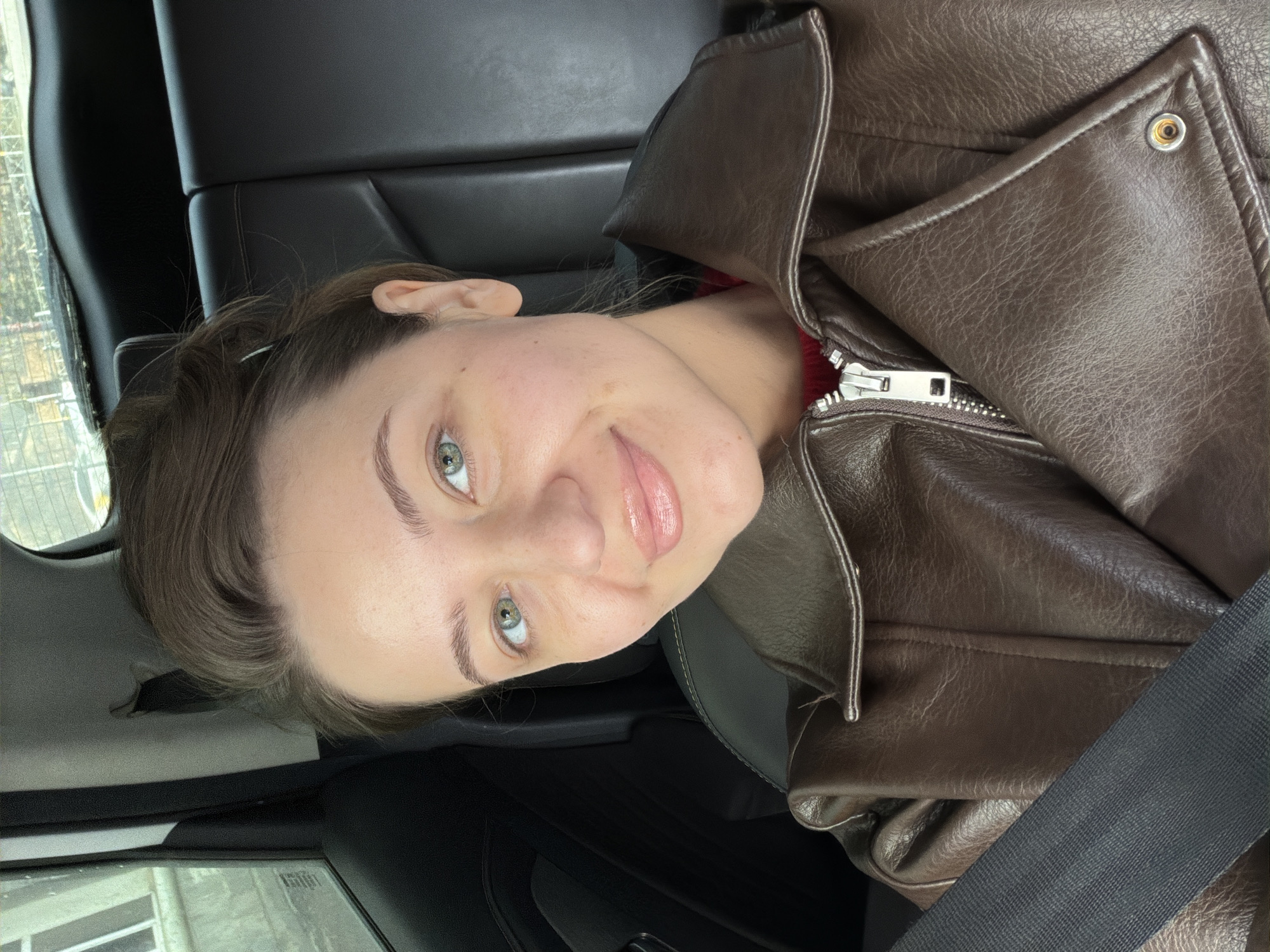 A young woman with fair skin and hazel eyes sits in the passenger seat of a car, likely in Kazan, Russia. She is wearing a brown leather jacket and a seatbelt is fastened across her chest. The car's interior is black leather. Outside the car window, there is a glimpse of buildings.