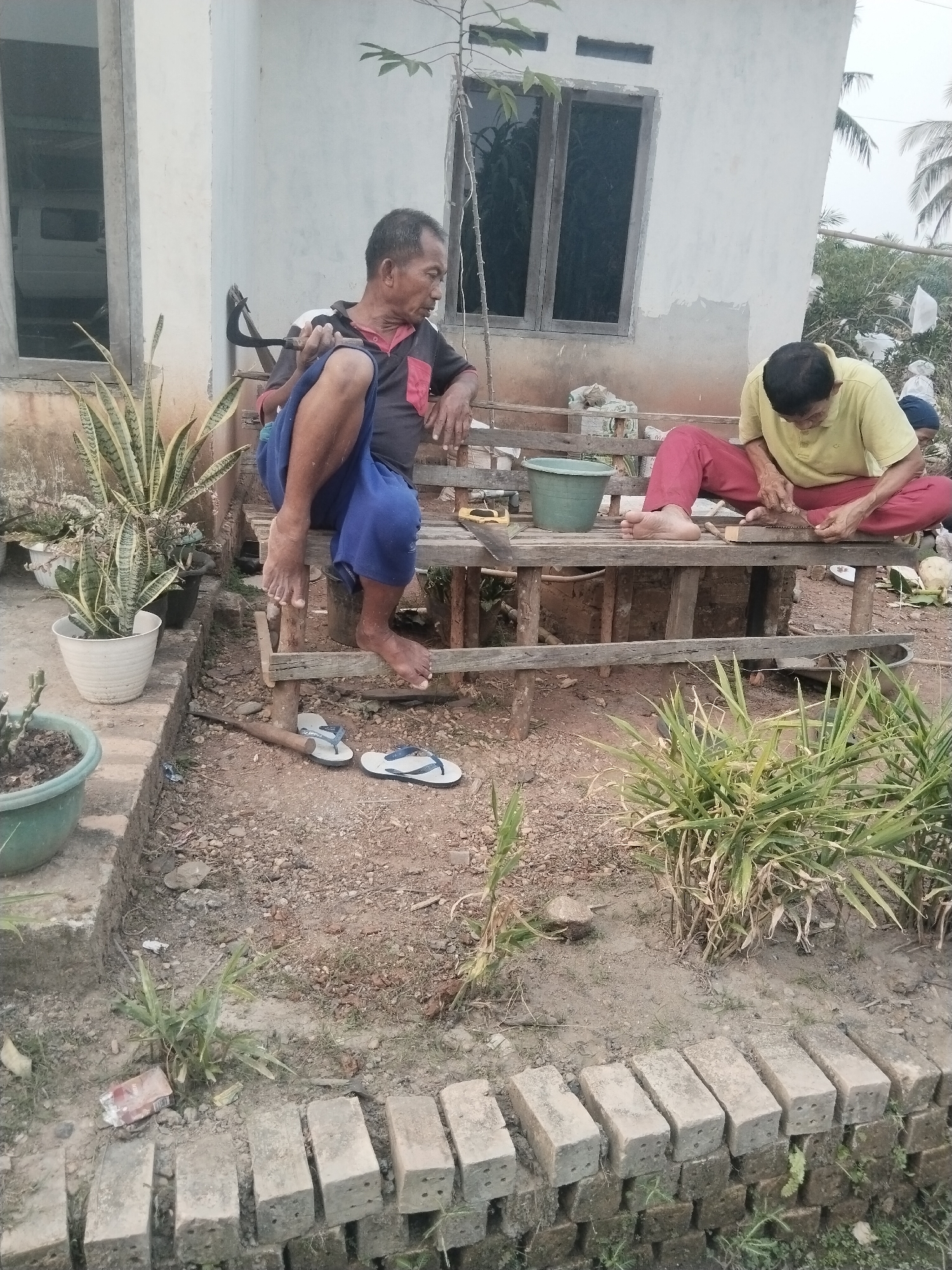 In Suka Ramai, Indonesia, two elderly Indonesian men are seated on a weathered wooden bench outside a simple, white-washed building with a dark-paned window. The man on the left, wearing a dark shirt with red accents and blue pants, is holding a curved metal tool, possibly a sickle, with his legs crossed. The man on the right, dressed in a yellow short-sleeved shirt and red pants, is focused on sharpening a wooden object with a knife. In front of the bench, a green bucket and a few other unidentifiable objects are present. The ground around the bench is covered with dirt, gravel, and sparse vegetation. A potted snake plant sits to the left of the bench, and further to the left, a larger green pot contains some plants. To the right of the bench, dense green foliage grows. A row of concrete bricks is laid out in the foreground. The overall scene depicts a moment of quiet activity in an outdoor, rural setting.