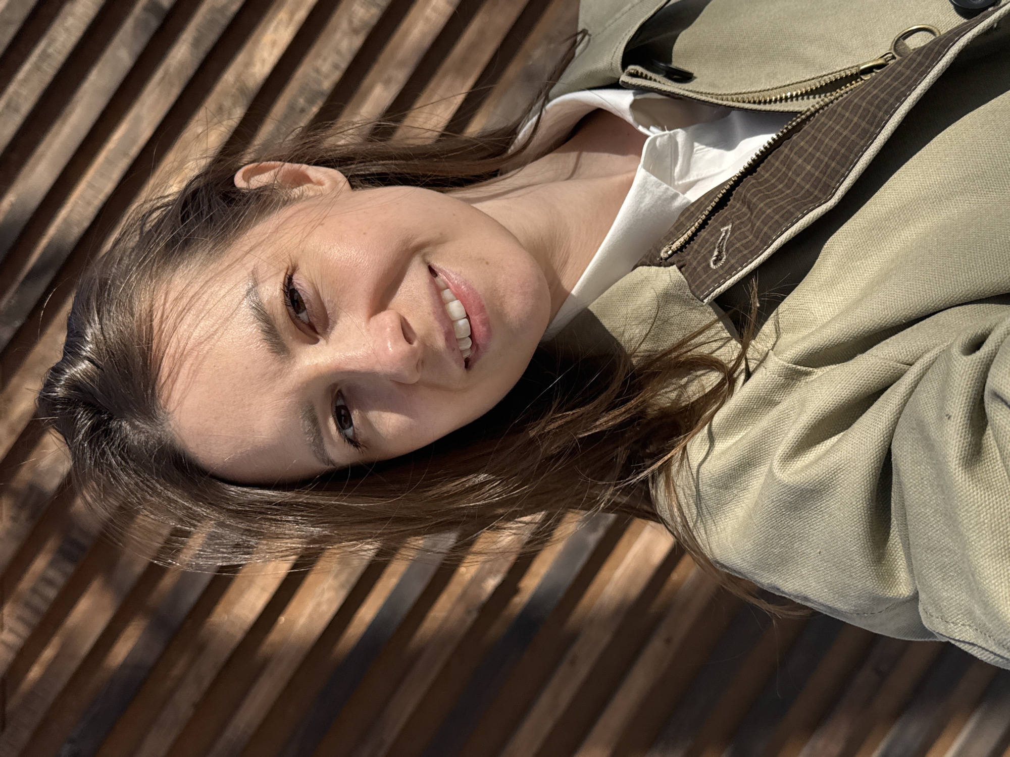 A woman is lying on a wooden bench in Chaikovsky, Russia. She has brown hair and fair skin, and she is smiling. She is wearing a white shirt and an olive green jacket. The bench is made of light brown wooden slats, and the sun is casting shadows across it.