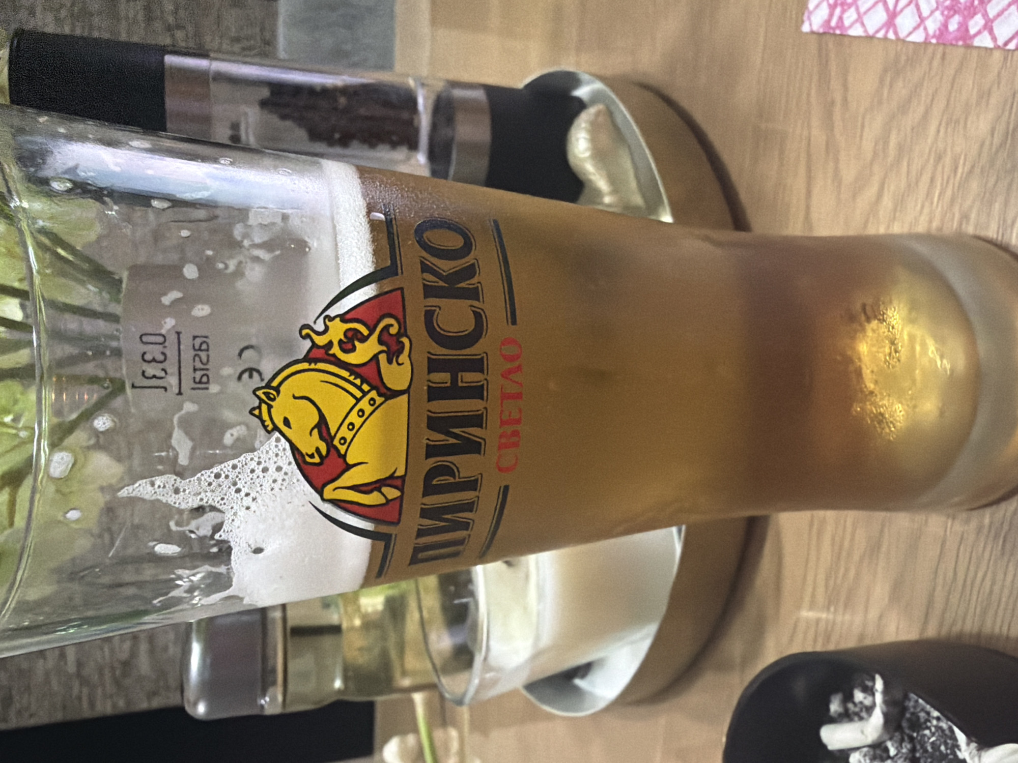 The image displays a close-up of a tall glass of beer on a light-colored wooden table, set in what appears to be an indoor restaurant or bar environment. The glass is filled with a golden-amber beer, topped with a substantial layer of white foam. Visible condensation on the glass indicates the beverage is cold.
The prominent label on the glass features the brand name "ПИРИНСКО" (Pirinško) in Cyrillic script, with "СВЕТЛО" (Svetlo), meaning "Light," written vertically underneath it, indicating a light lager. The logo associated with the brand is a stylized yellow horse on a red background, contained within a shield-like shape. Additionally, embossed on the glass are the volume markers "0.33 L" and a "CE" mark, along with some indistinct alphanumeric characters.
In the background, a pepper grinder with a black cap and a clear body filled with black peppercorns is visible. Parts of other objects, including a metallic element and a small white ceramic dish, are also discernible. In the bottom right corner, a dark, possibly black, small container is partially visible, potentially an ashtray or condiment holder. A corner of a pink and white patterned napkin or paper is seen in the upper right.
No people are present in the image, and no specific event is taking place, other than the beer being served for consumption. The scene suggests a relaxed atmosphere. The Bulgarian beer brand "Pirinško" aligns with the location context of Shumen, Bulgaria. The time of day and weather conditions are not ascertainable from the indoor setting.