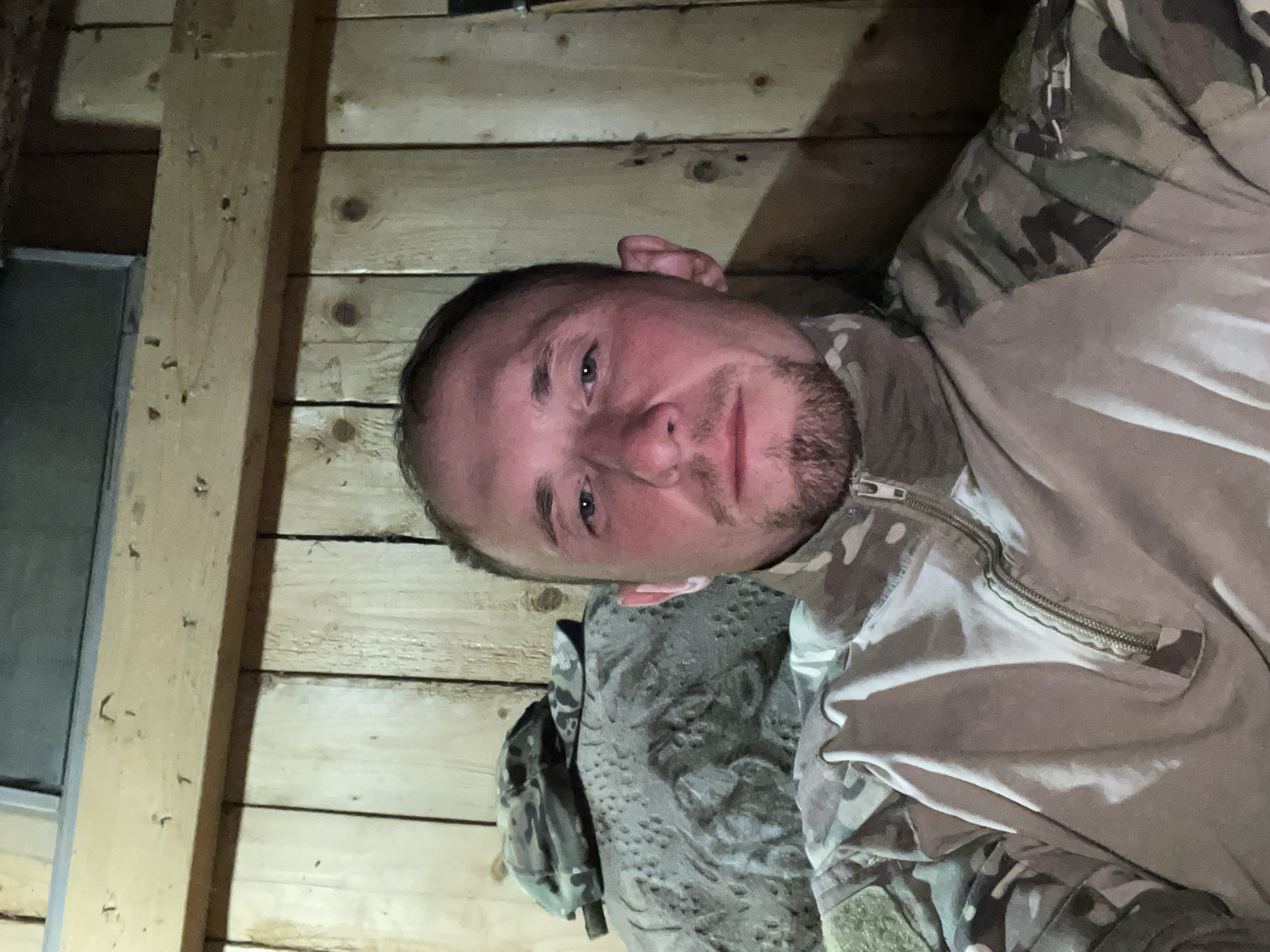 A selfie of a man with light skin and a short beard, wearing a camouflage uniform, is taken indoors. He has short brown hair and blue eyes, with a slight blush on his cheeks. The background consists of wooden planks, suggesting an interior of a rustic structure. To his left, a camouflage cap rests on a textured, possibly rocky or fabric surface. The lighting appears to be artificial and dim. The man is looking directly at the camera.