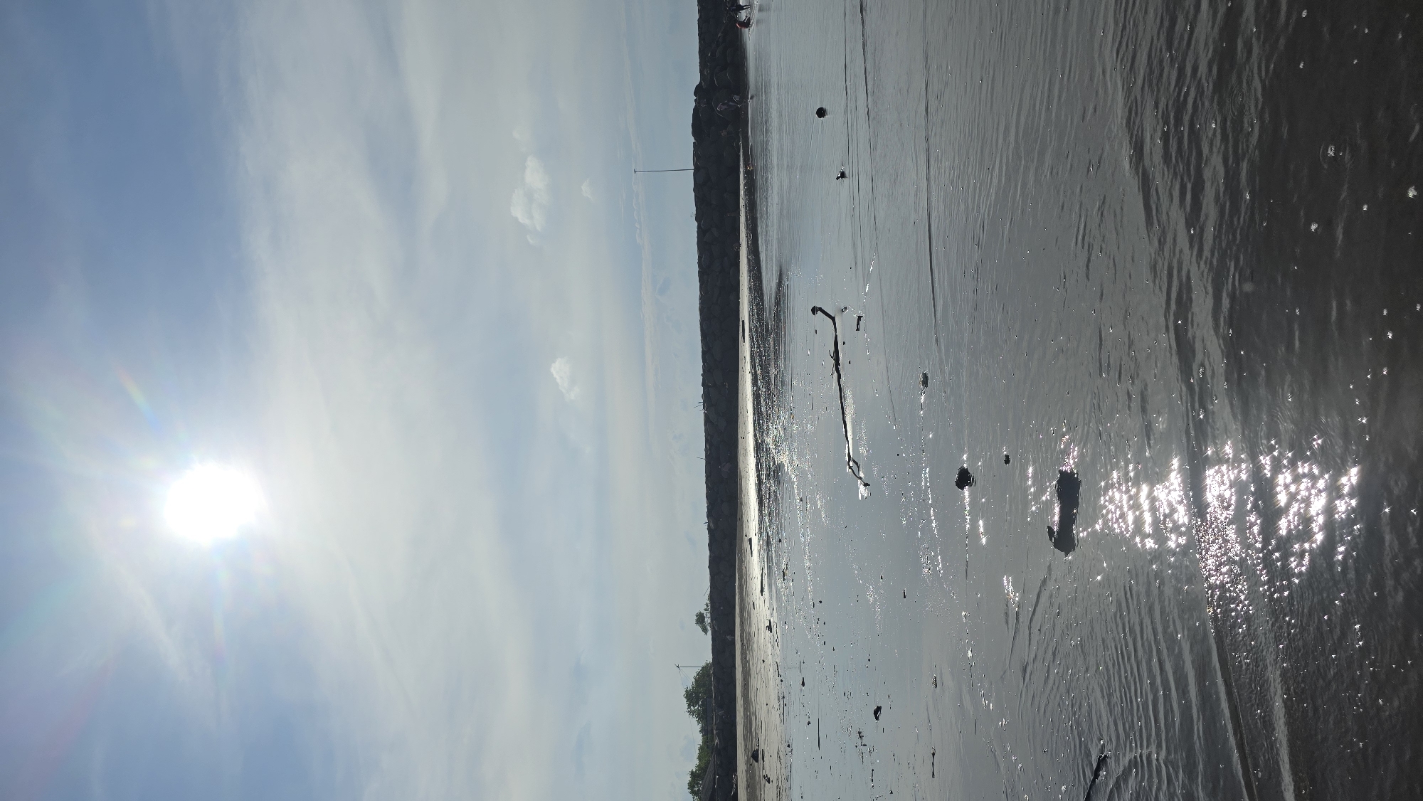 The image captures a coastal scene at Geulumpangsulu Timu, Indonesia. The foreground is dominated by a wet, reflective beach or mudflat. Scattered debris, including a notable branch, lies on the surface. The water reflects the sunlight, creating sparkling highlights. A dark embankment forms a line separating the beach from the land or built environment. The sky features a bright sun partially obscured by clouds, with a blue hue visible around it. Buildings or structures are faintly discernible in the distance along the embankment.
