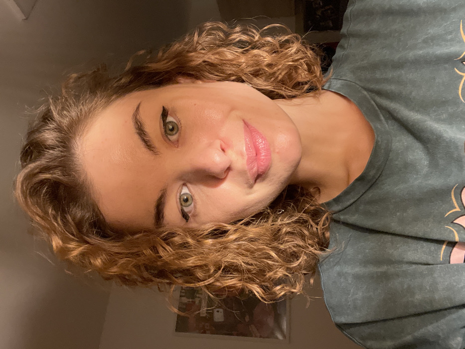 A close-up, frontal portrait features a female subject in an indoor setting. The subject has light to medium skin, light green eyes with black winged eyeliner, and shoulder-length, coppery-brown curly hair. Her lips are closed in a subtle smile, enhanced with a glossy pink lip product. She is wearing a dark green, possibly acid-washed, t-shirt with a partially visible light pink graphic design on the lower chest. The background is dimly lit, with a framed item, possibly a picture or poster, visible on the wall to the upper left. A direct light source illuminates the subject from the front-right. The precise location within Boreti, Montenegro, is not discernible from the visual information provided.
