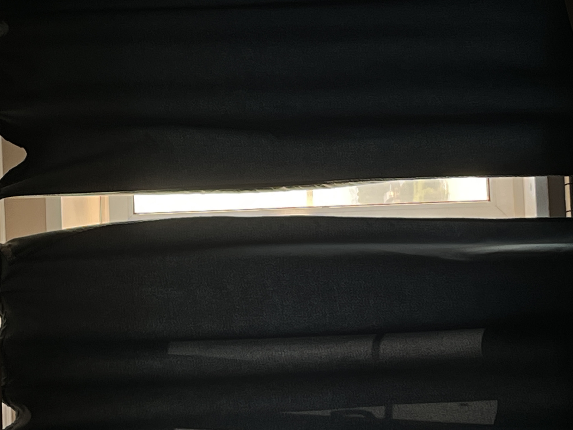 The image shows two dark curtains partially drawn back, revealing a sliver of a window. The curtains appear to be made of a textured, heavy fabric and are a dark color, possibly black or deep navy. Sunlight is filtering through the gap in the curtains, illuminating the edges of the fabric. Through the window, a blurry view of what appears to be an outdoor scene can be seen, with hints of greenery and possibly some buildings in the distance. The overall impression is of a dimly lit interior with a bright exterior partially visible. There are no people or specific objects clearly identifiable in the scene.