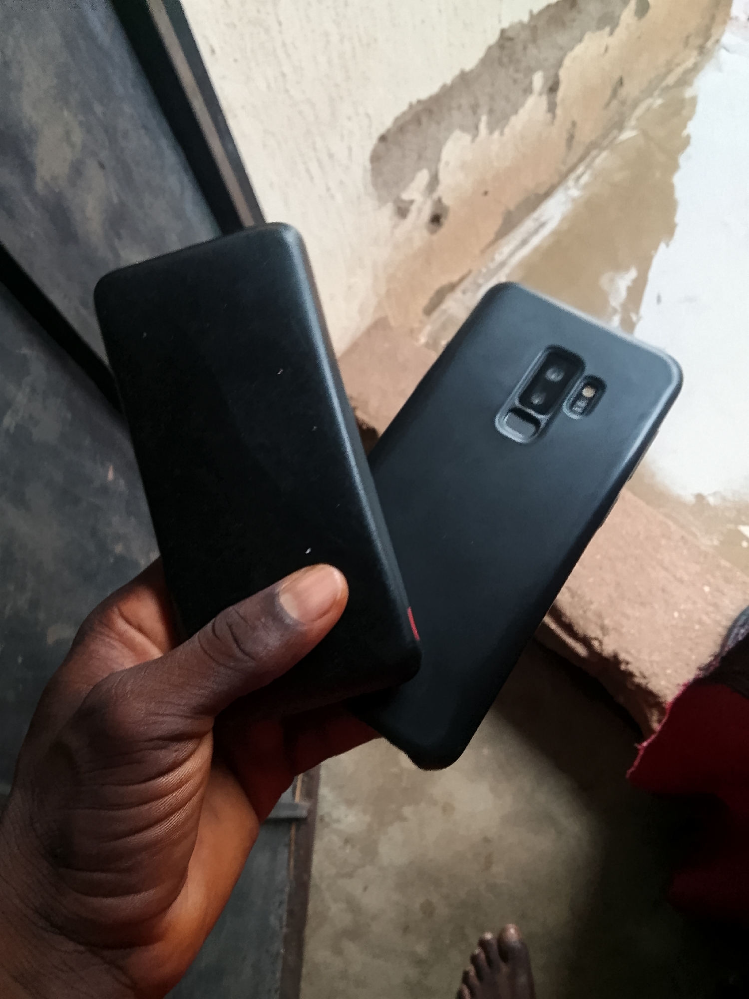 In Jalingo, Nigeria, a dark-skinned hand holds two black smartphones. One phone, on the left, is rectangular with rounded corners. The second phone, on the right, is sleeker with a camera module on the back featuring two lenses and a flash. The background shows a textured wall with peeling paint and a floor that appears to be made of concrete or a similar material. A bare foot is visible in the lower right corner of the image.