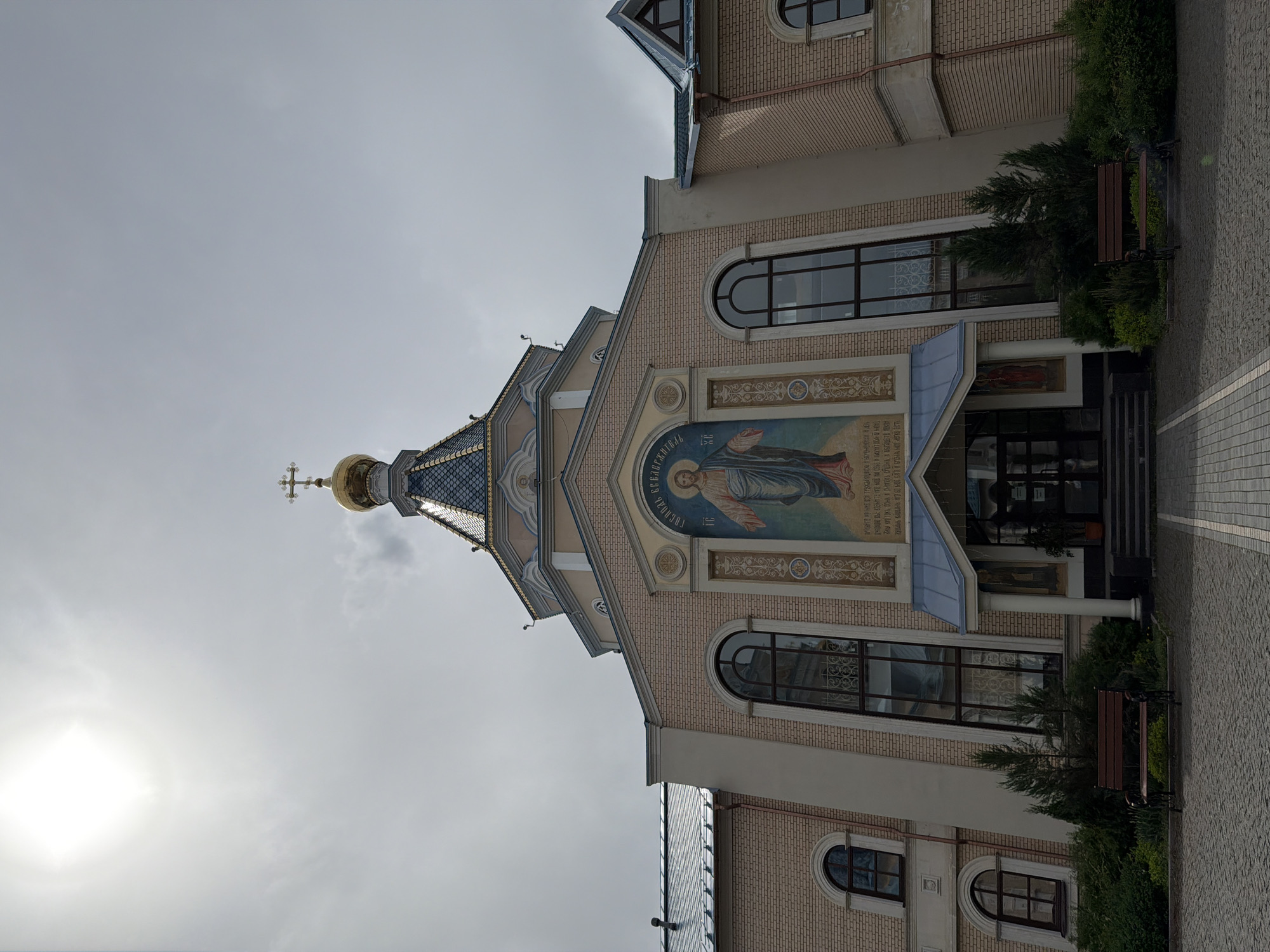 This is an image of the facade of the Orthodox St. Michael's Cathedral in Izhevsk, Russia. The image has been rotated to a vertical orientation. The church is constructed of brick and features multiple windows with rounded tops. A mural of Jesus Christ is displayed above the entrance. A gold dome with a cross rises above the building. Benches sit on either side of the entrance, nestled amongst greenery. The sky is overcast.