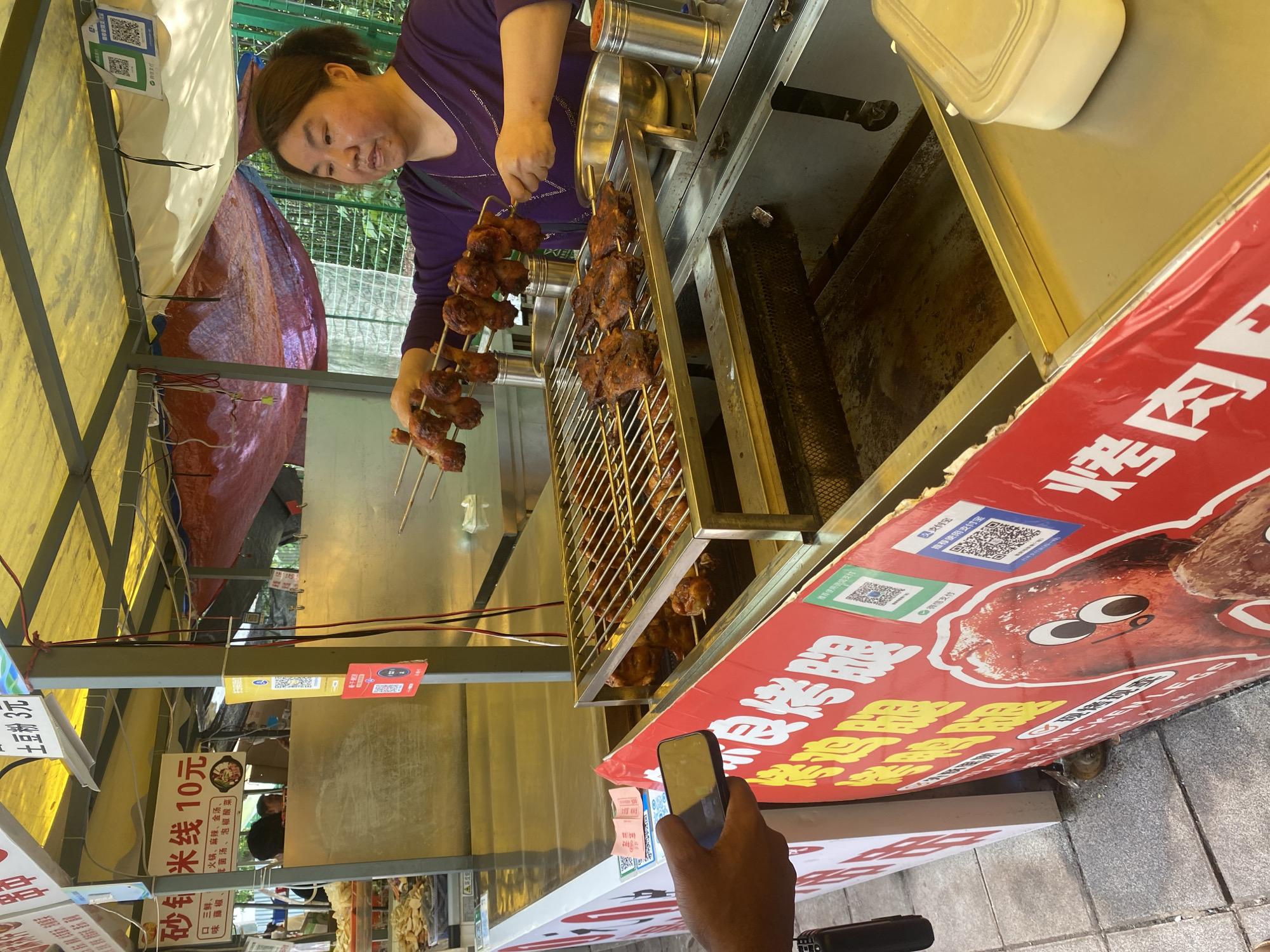 The scene depicts a food stall in Shapingba District, China. A woman wearing a purple shirt is tending to skewers of cooked chicken legs placed on a metal rack. In front of the stall is a red sign featuring a cartoonish depiction of a chicken leg and the Chinese characters "烤肉", which means "grilled meat", as well as the English text "CHICKEN LEGS". The stall also displays pricing signs; one reads "米线 10 元" (rice noodles 10 yuan), another "土豆粉 3元" (potato noodles 3 yuan). A hand holding a smartphone is visible in the lower-center of the image, possibly taking a picture or scanning a QR code. The environment is a typical outdoor market setting.