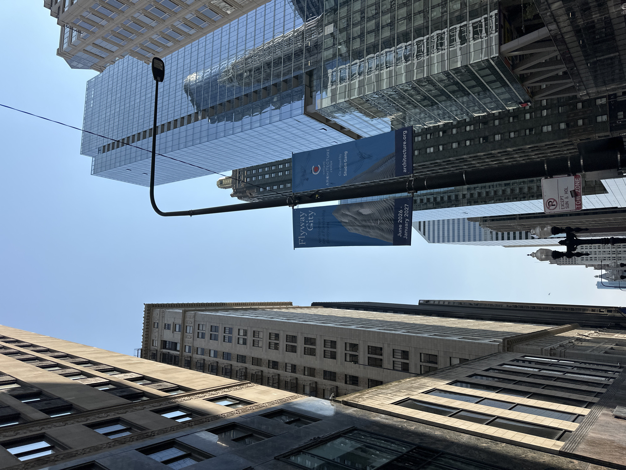 This is a low-angle, upward-facing outdoor shot of a city street in Chicago, Illinois, USA, under a clear blue sky. The image is dominated by several tall buildings with distinct architectural styles. On the left, a dark-colored, modern building with many windows is visible, angled towards the viewer. To its right, a lighter-colored, older building with a decorative facade and rows of small windows stands. Further to the right, a sleek, glass-covered skyscraper reflects the sky and surrounding buildings. Two blue banners are hanging from a lamppost in the center of the frame. The left banner reads "Flyway City" and lists dates "June 2026 - January 2027." The right banner displays the "Chicago Architecture Center" logo and states "Co-curated by Studio Gang" and "architecture.org". In the lower right, a "R EXCEPT SUN & HOL TOW ZONE" sign is attached to a lamppost. The scene suggests an urban environment, possibly a downtown area, with a focus on architecture and potentially an upcoming event.