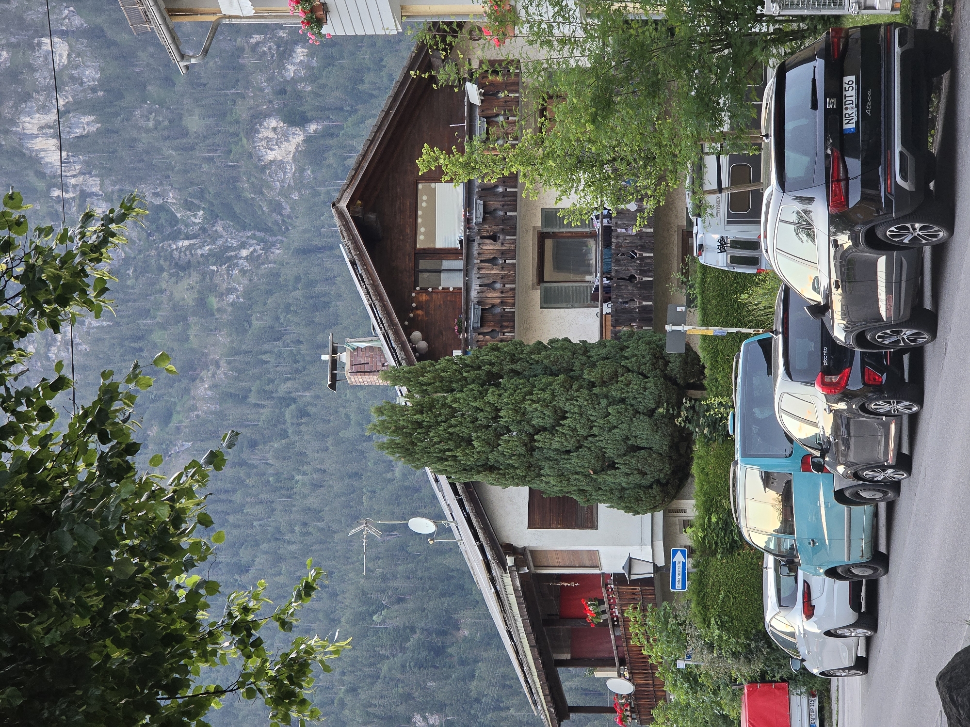 A street scene in Garmisch-Partenkirchen, Germany, features a row of parked cars on the right side of the image. From left to right, a white hatchback, a teal van, and a black SUV are visible. The black SUV has a license plate that reads "NR DT 56". Behind the cars, a multi-story building with balconies and wooden architectural details is situated. The building is surrounded by lush green trees and foliage. In the background, a steep, forested mountain slope rises. The sky is not visible due to the framing of the image.