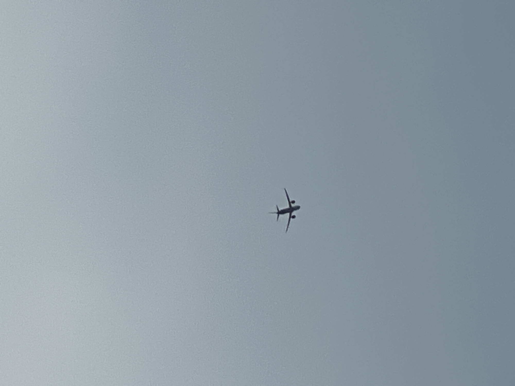 A passenger airplane is captured in flight against a uniformly gray, overcast sky. The aircraft appears as a dark silhouette with clearly defined wings, fuselage, and tail. The airplane is positioned in the lower center of the frame, flying horizontally. The sky is featureless and appears to be the only environmental element present. There are no discernible landmarks or geographical features visible, making it impossible to determine the specific location in Iserlohn, Germany, or any other city.