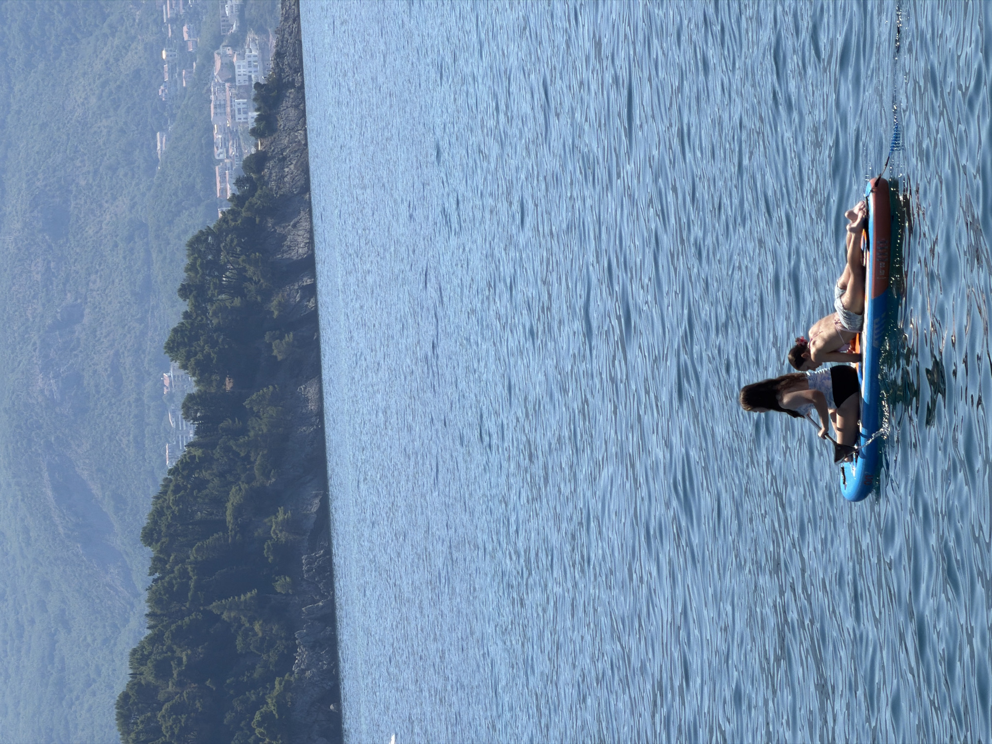 Two individuals are on a paddleboard in the blue, wavy sea, with the coast of Budva, Montenegro, in the background. The person in the foreground is a young female with long brown hair, wearing a white and black patterned top and black shorts. She is sitting on the paddleboard and holding a paddle. The person behind her is a young female wearing a patterned bikini top and patterned shorts. She is lying on her stomach on the paddleboard, facing away from the camera. The paddleboard is blue and orange. In the background, there is a rocky coastline covered in green trees and some buildings. The sea is calm with gentle ripples. The sky is clear and blue.