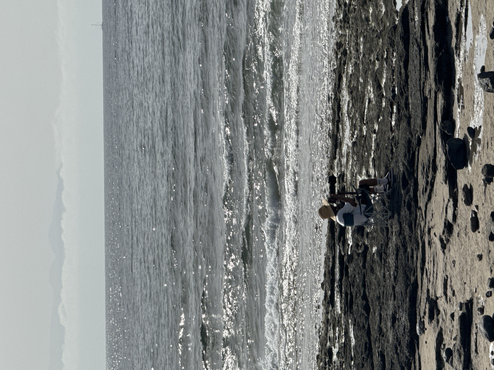 A lone individual sits on a foldable chair on a rocky shoreline, looking out at the ocean. The person is wearing a straw hat, a white t-shirt with blue accents, dark shorts, and white socks with sneakers. They are holding a camera and appear to be taking pictures. The ocean is calm with gentle waves, and the sun glints off the water. In the distance, a sailboat is visible on the horizon, and a misty mountain range can be seen. The foreground consists of dark, jagged rocks and sandy patches. The overall scene is one of peaceful solitude in a coastal environment. The location is identified as Arona, Spain, a municipality on the southern coast of Tenerife.