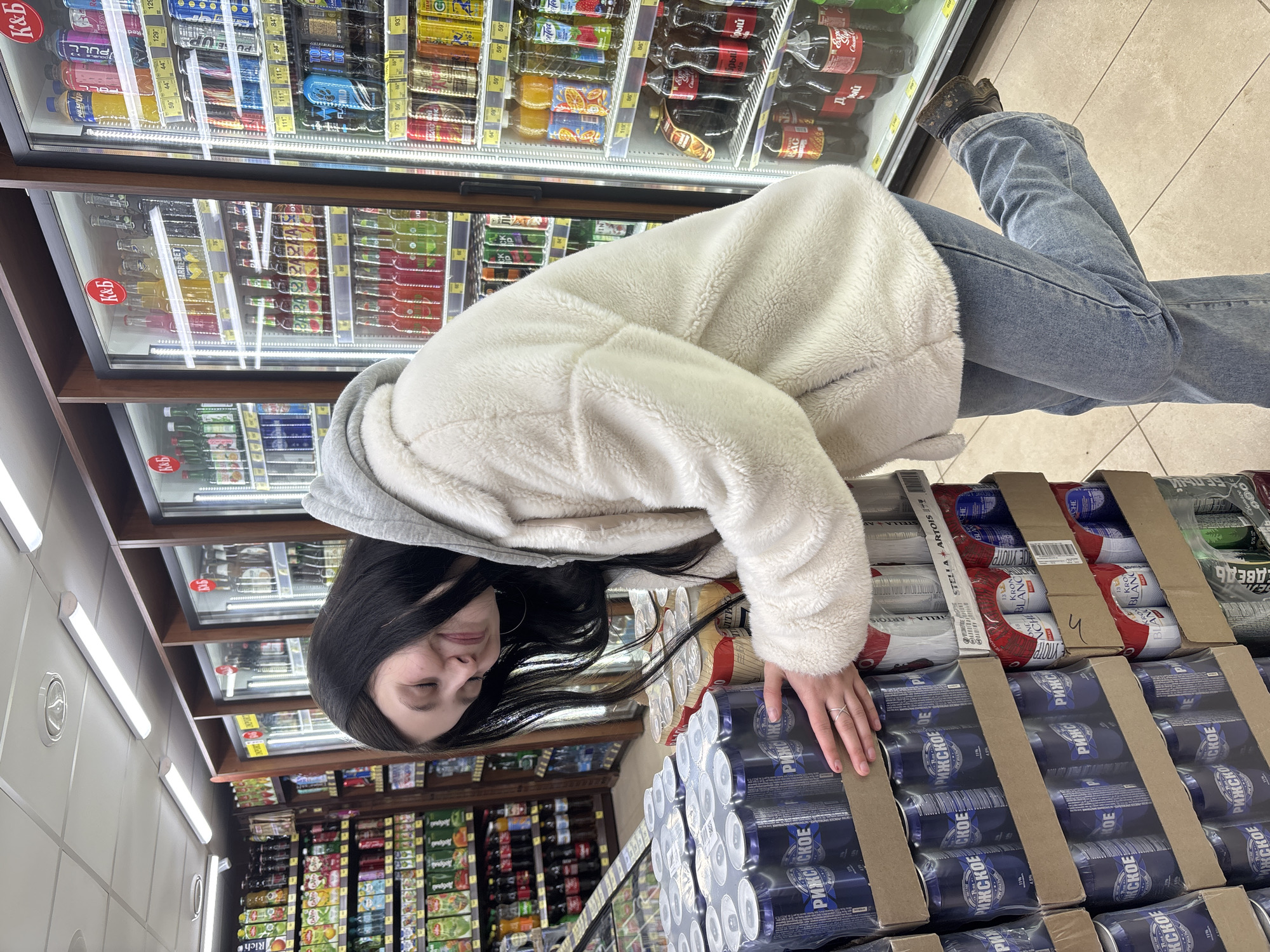 The image shows a person standing in a store aisle, likely in Фоки, Russia, based on the Cyrillic labels on the products. The individual is wearing jeans, shoes, and a cream-colored fuzzy jacket with a gray hood. They appear to be browsing the beverage section. The shelves behind them are stocked with a variety of canned and bottled drinks, including brands like Stella Artois and РИЖСКОЕ. Several price tags are visible. They have their hand on packs of canned beverages.