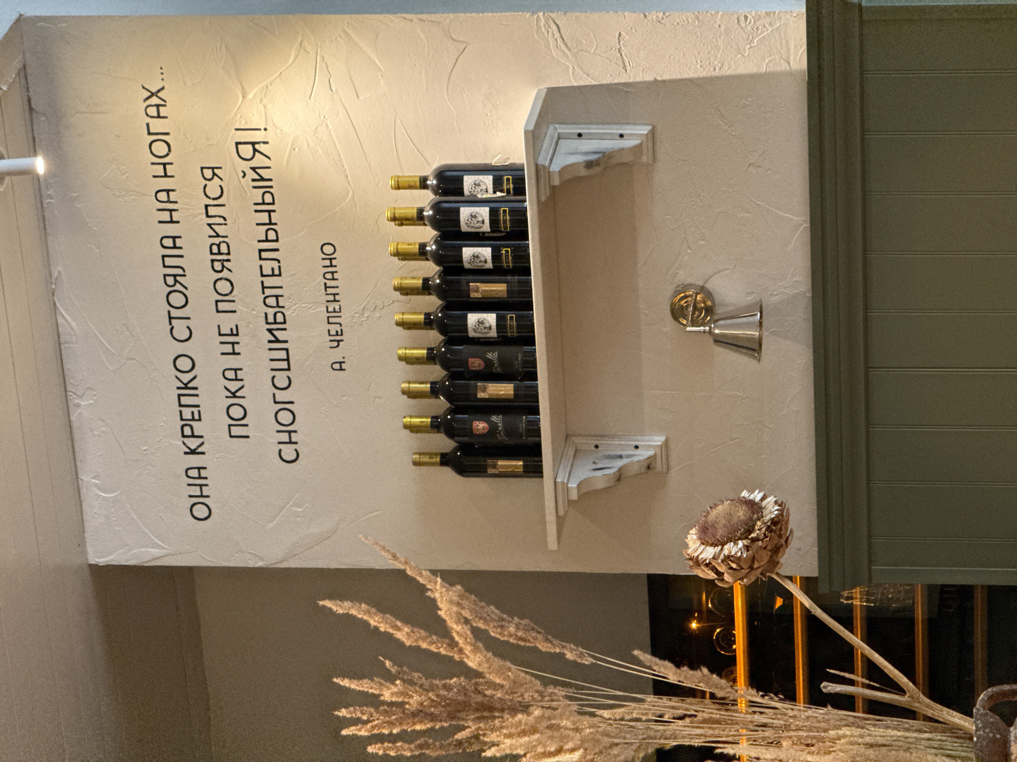 This image shows a vertical display in what appears to be an indoor setting, likely a restaurant or wine bar in Krasnoyarsk, Russia. On the left side of the display, there is white wall texture and Russian text, "ОНА КРЕПКО СТОЯЛА НА НОГАХ... ПОКА НЕ ПОЯВИЛСЯ СНОГСШИБАТЕЛЬНЫЙЯ! A. ЧЕЛЕНТАНО", which translates to "SHE STOOD FIRMLY ON HER FEET... UNTIL STUNNING ME APPEARED! A. CHELENTANO."
To the right of the text, a shelf constructed of painted medium density fibreboard contains 8 bottles of red wine. The wine labels are black with gold letters and display the name "Prinelli". The wine bottles are staggered between two horizontal supports.
To the right of the bottles is a faux silver-plated wall mounted candle holder.
In the foreground is a dried flower arrangement consisting of beige pampas grass and a dried artichoke. In the background is a window with metal bars. The setting suggests a sophisticated and perhaps humorous presentation related to wine.