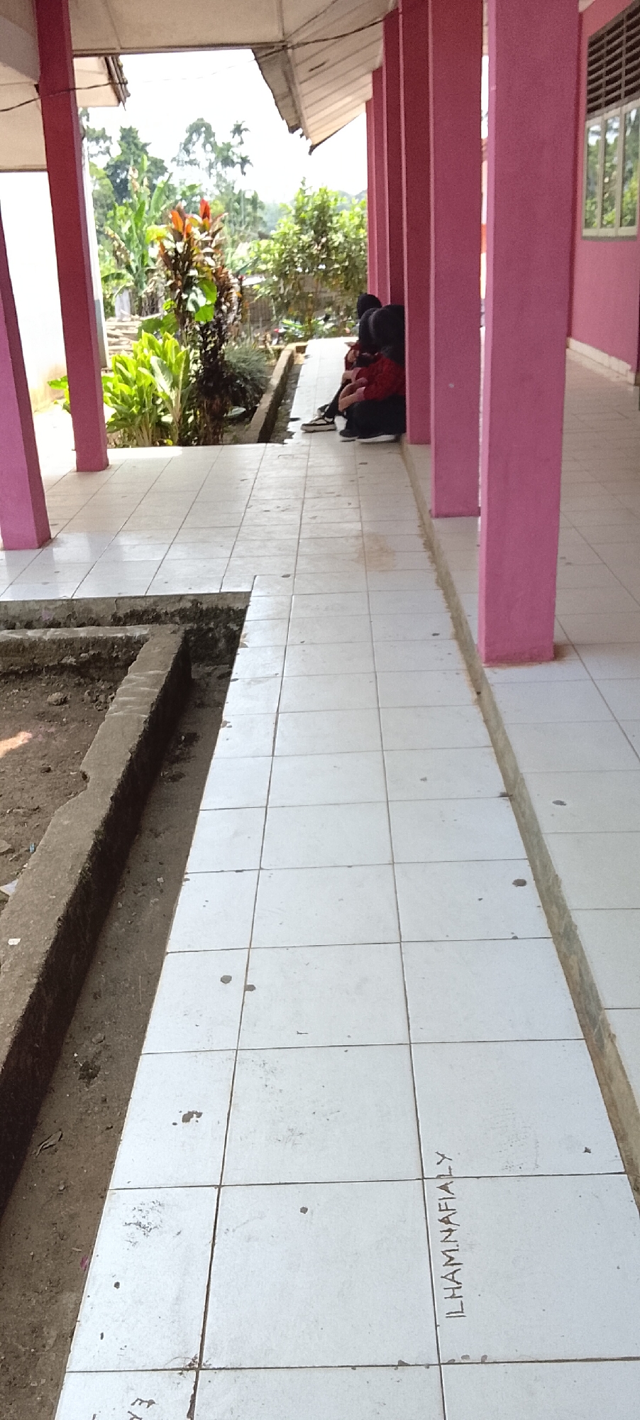 A photograph taken from a low angle shows a row of pink pillars supporting a covered walkway. The walkway is tiled with white square tiles, some of which are dirty and stained. To the left of the walkway, there is a raised planter box filled with soil and some vegetation, including a plant with red and green leaves. Further down the walkway, two individuals are seated, facing away from the camera. They are wearing dark clothing, and one of them appears to be wearing a red top. A window with green frames is visible on a pink wall in the background. The scene appears to be outdoors, with trees and foliage visible beyond the walkway. The words "ILHAM.NAFAALY" are written in dark marker on the tiled floor in the foreground.