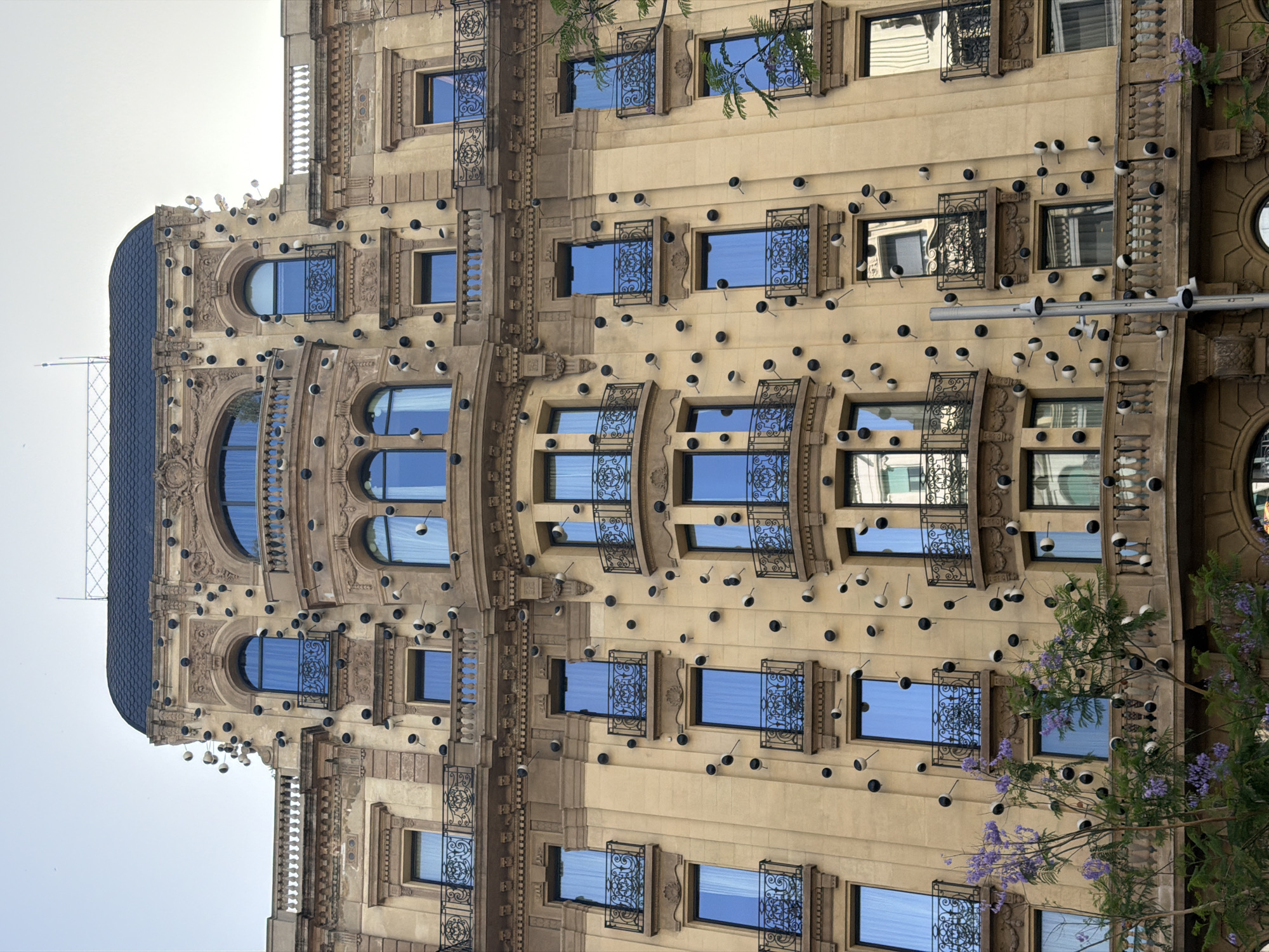 The image shows a facade of a multi-story building, likely in Barcelona, Spain, due to the architectural style and presence of jacaranda trees with purple flowers. The building is adorned with numerous black and white spherical objects attached to its exterior walls and balconies. These objects appear to be decorative or artistic installations. The building itself is classical in style, with ornate stonework, arched windows, and balconies with wrought-iron railings. The sky above is overcast. The lower part of the image shows more of the jacaranda trees in bloom, obscuring some of the lower levels of the building. No people are visible in the image.