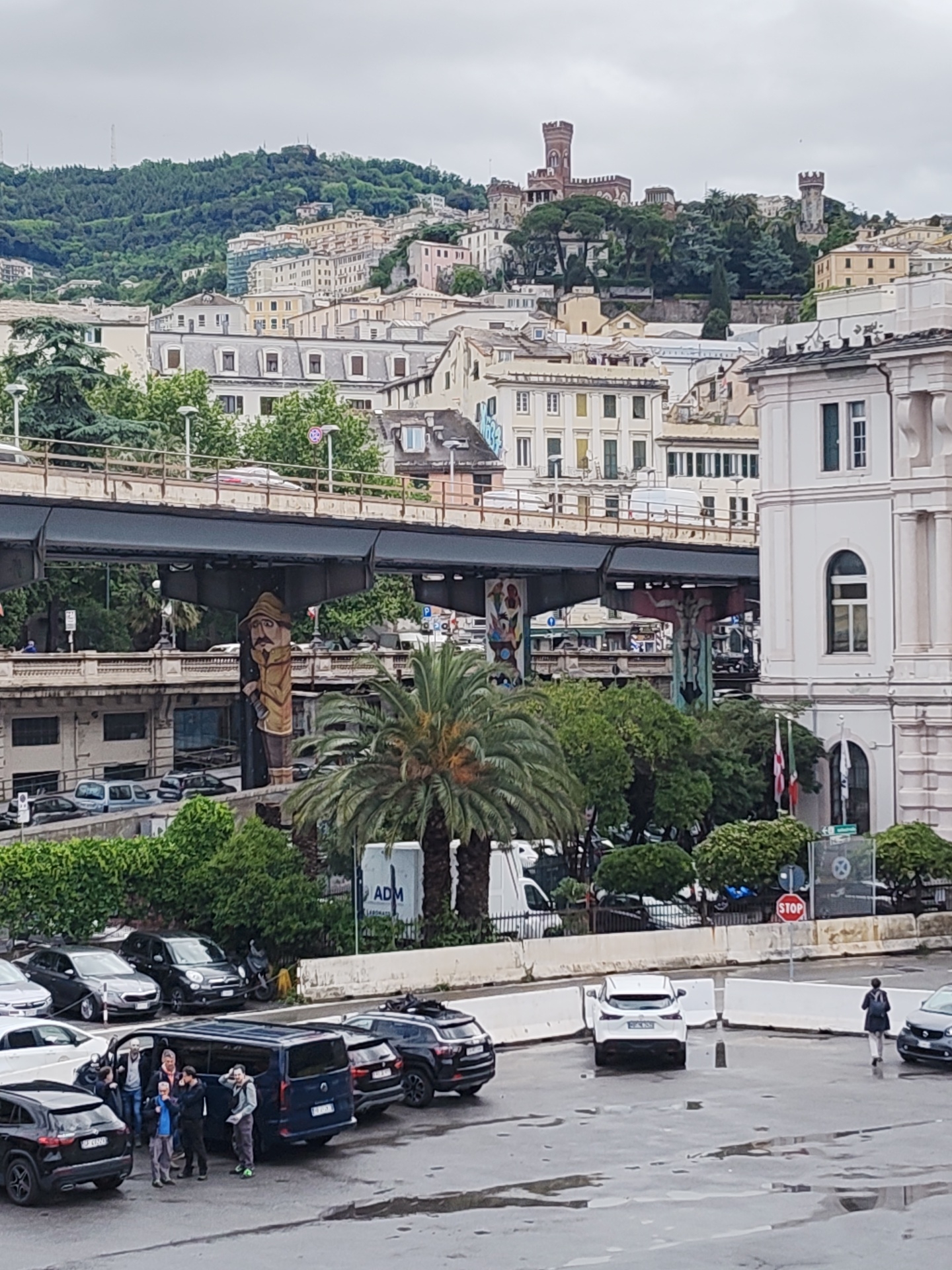 The image depicts a scene in Genoa, Italy, with a cityscape backdrop and a foreground parking area. In the background, a castle or fortress structure stands atop a green hillside amidst residential and commercial buildings. An elevated roadway, potentially a highway or bridge, cuts across the middle ground of the scene, with support pillars adorned with art or murals. In the parking area, several vehicles, including cars and a van, are visible. A group of approximately six people is gathered around one of the vehicles, seemingly interacting. Another individual is walking across the parking area, away from the camera. A palm tree and other greenery are interspersed throughout the scene, particularly between the elevated roadway and the parking area. A building with architectural details, including arched windows and columns, is visible on the right side of the image, with flags displayed in front. There are parking enforcement signage and a stop sign in the foreground, near the parking lot. The weather appears overcast, with a grey sky and wet pavement.