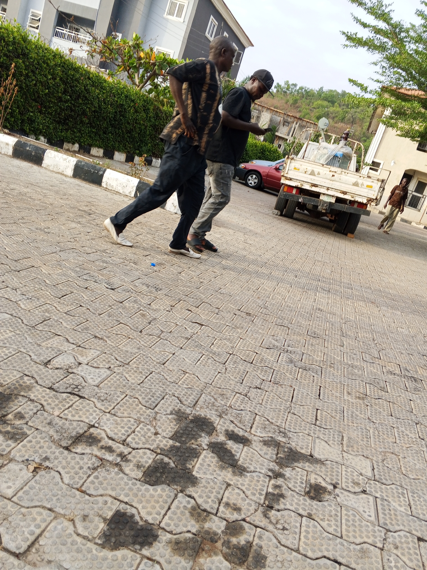 The image is taken outdoors on a paved surface of interlocking concrete pavers. Two men are walking, one wearing a patterned shirt and dark pants, the other in a black shirt, gray jeans, and a baseball cap. A white truck is parked nearby, with a red car partially visible behind it. Another man is walking in the background, further down the road. In the background there is a residential building and a hedge. The location might be a residential area in the city of Lokoja, Nigeria.
