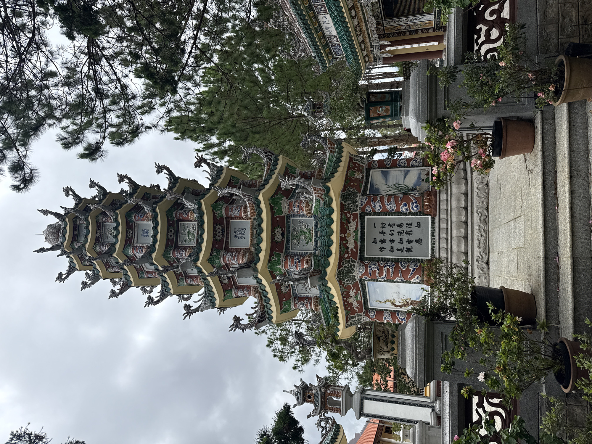 The image depicts the Thien Vuong Pagoda, a prominent Buddhist temple located in the city of Da Lat, Vietnam. The pagoda, a multi-tiered structure adorned with intricate dragon motifs and colorful ceramic tiles, stands tall against an overcast sky. Pine trees frame the scene, their branches extending into the frame. In the foreground, potted plants and stone steps lead up to the pagoda's base, where decorative panels and inscriptions are visible. To the left and right, other temple structures, characterized by their green tiled roofs and ornate detailing, are partially visible. The overall impression is one of a serene and architecturally rich religious site amidst a natural landscape.