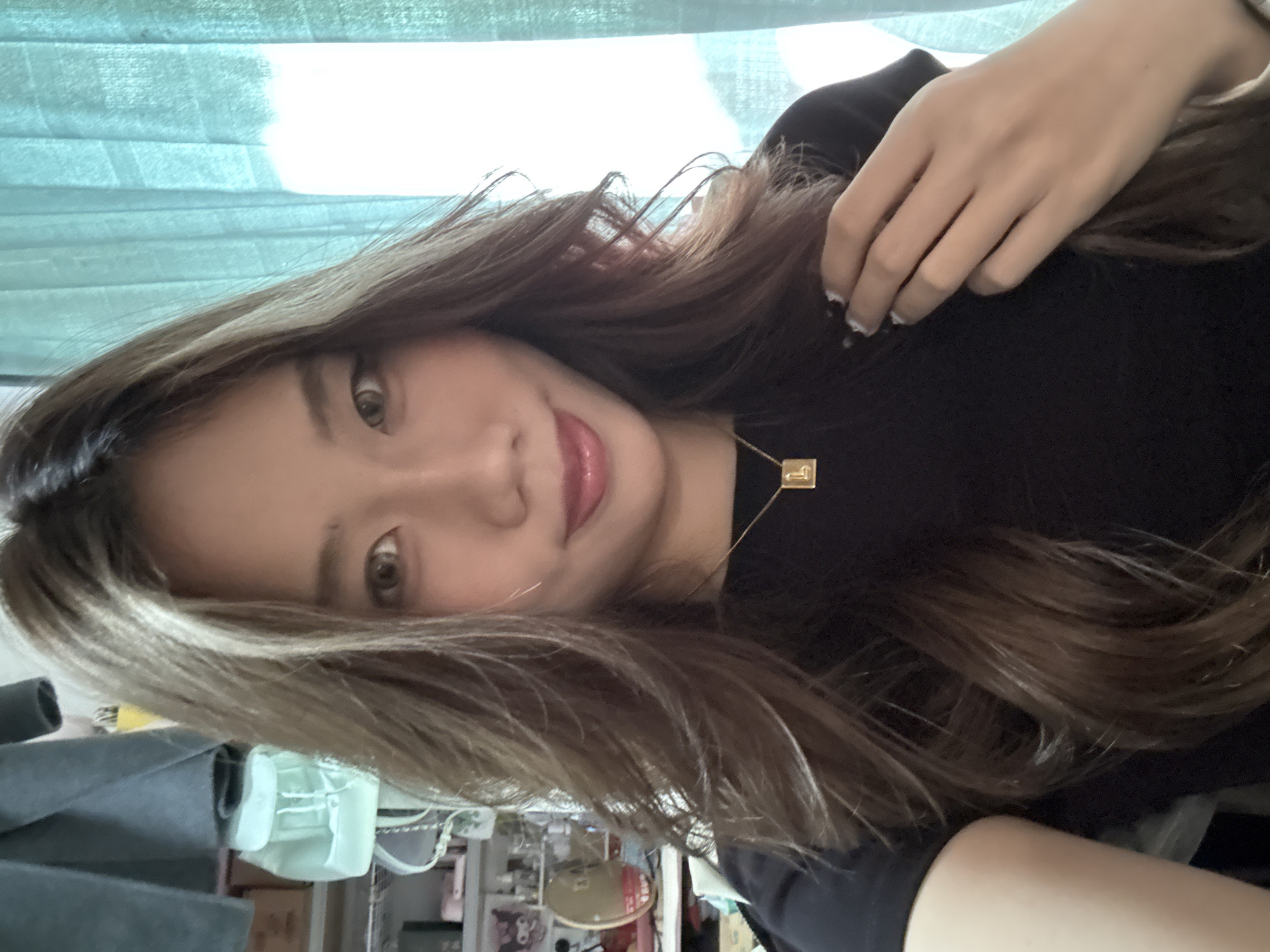A woman is pictured in a selfie. She has long, brown, wavy hair, and fair skin. She is wearing a black top. A gold necklace is visible on her chest. Her nails are painted with white tips. The background appears to be a room with various items, including what looks like a mask, possibly indicating an indoor environment. The image orientation is rotated. No specific details suggest a location in the city of Nan'an District, China.