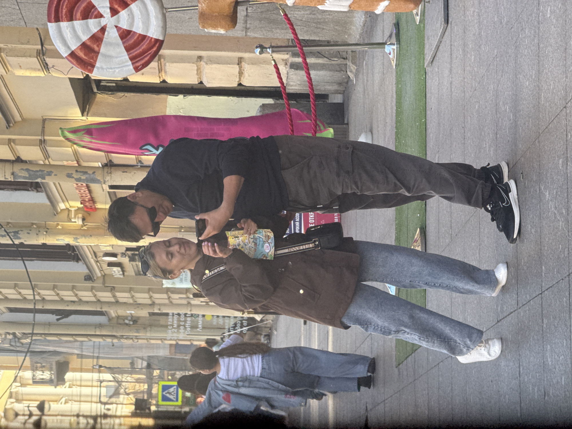 This image captures a street scene in Saint Petersburg, Russia. Two individuals are the primary subjects, a man and a woman, seen from the waist down, interacting with their phones. The man is wearing dark pants and black sneakers, while the woman has on a brown jacket and blue jeans, paired with white sneakers. They appear to be standing on a paved sidewalk, with a green strip of what looks like artificial turf to their left. In the background, a large, round, red and white object, resembling a lollipop or a decorative target, hangs overhead. There are also some pink and green surfboards leaning against a building, suggesting a possible surf shop or theme. The overall impression is of a casual, possibly tourist-oriented area. The lighting suggests it is daytime, with a bright, diffused light, perhaps indicating an overcast day or late afternoon. Visible text in the image includes "Coffee to", "Лимон" (Lemon), "POLO ASSN", and "SPOLO AS", likely storefront signs or product labels. The mood appears relaxed, with the focus on the individuals and their technology.