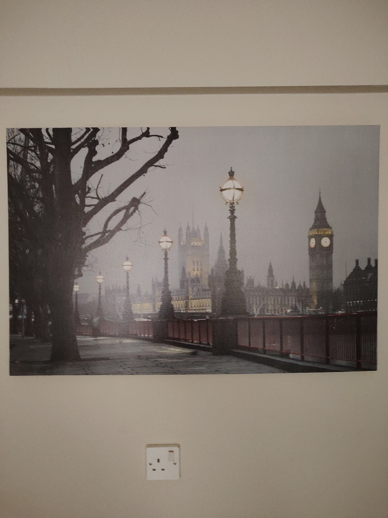 The image displays a misty urban landscape, likely captured at dusk or night, in the city of Huntingdonshire, United Kingdom. In the left foreground, a large, bare deciduous tree extends its dark, gnarled branches across the upper left quadrant. A paved walkway or street extends from the lower left, running parallel to a dark, sturdy railing or balustrade that features a red top edge, defining the right side of the immediate foreground. Along this railing, several ornate streetlights are positioned at intervals, each emitting a warm, circular glow, illuminating the ambient fog. In the mid-ground, indistinct architectural forms are partially obscured by the atmospheric mist. The background features several prominent gothic-style buildings, their illuminated windows and intricate details faintly discernible through the haze. A tall, dark clock tower stands distinctly on the far right, its two visible clock faces brightly lit, marking time in the atmospheric scene. No people are visible in the depiction.