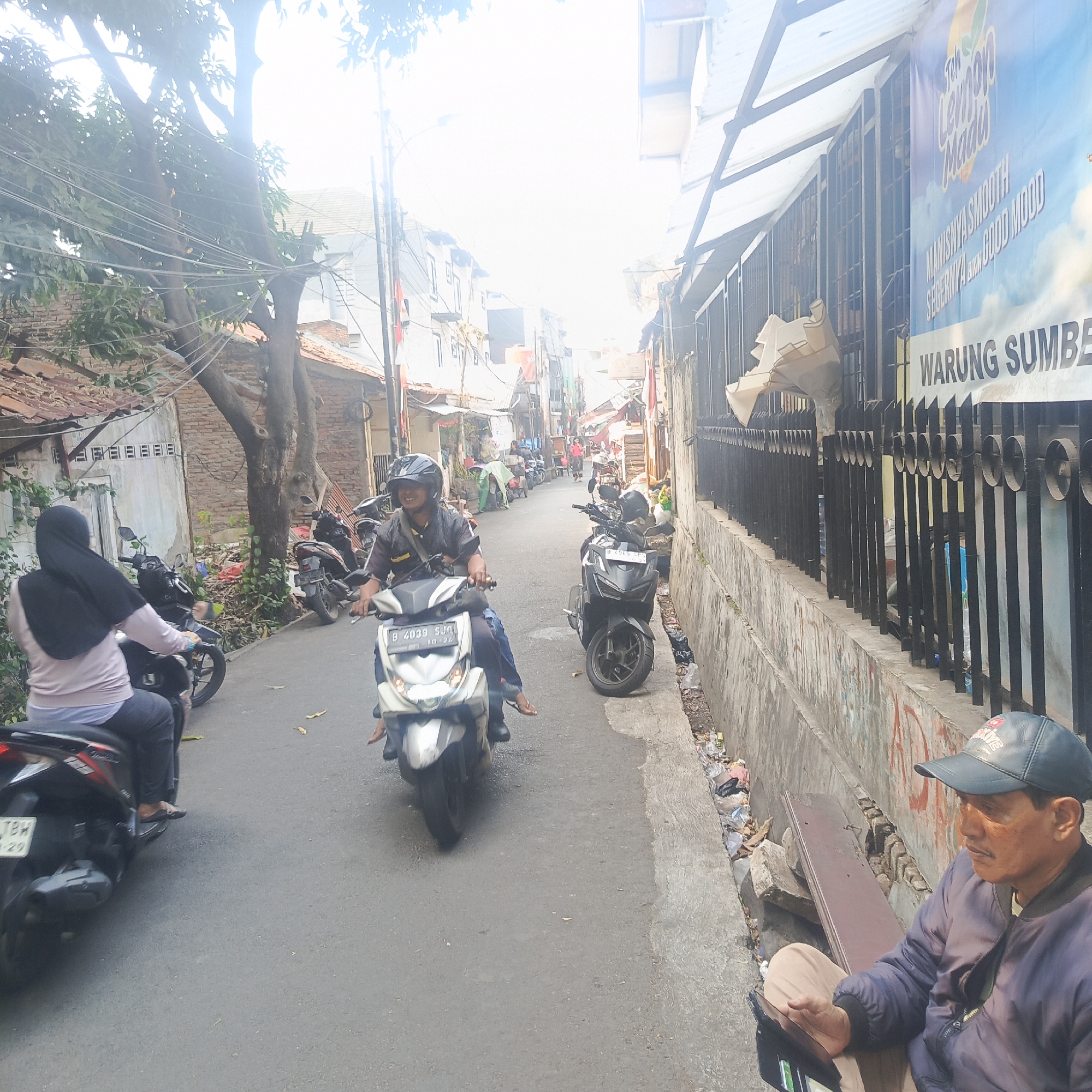 The image depicts a narrow street in South Jakarta, Indonesia, likely a residential or commercial alley. The scene is busy with multiple motorcycles, some in motion and others parked. In the foreground, a person wearing a black hijab and a light-colored shirt rides a black motorcycle. Further down the street, two men are on a white motorcycle, one driving and the other a passenger, both wearing helmets. Another black motorcycle is parked on the right side of the street, and more motorcycles are visible further in the background, suggesting a bustling local area. The buildings lining the street are mostly two-story structures with tiled roofs and some are covered by corrugated metal awnings. A banner with text that includes "WARUNG SUMBER" and "Teh Lemon Madu" is visible on the right side of the street, indicating a local food or beverage stall. The environment appears somewhat cluttered, with debris and some overgrown vegetation near the buildings and along the drainage ditch on the right. A man in a dark jacket and a baseball cap is seated on a bench on the right side of the image, looking down at a tablet. The overall atmosphere suggests a typical day in a densely populated urban area in Indonesia.