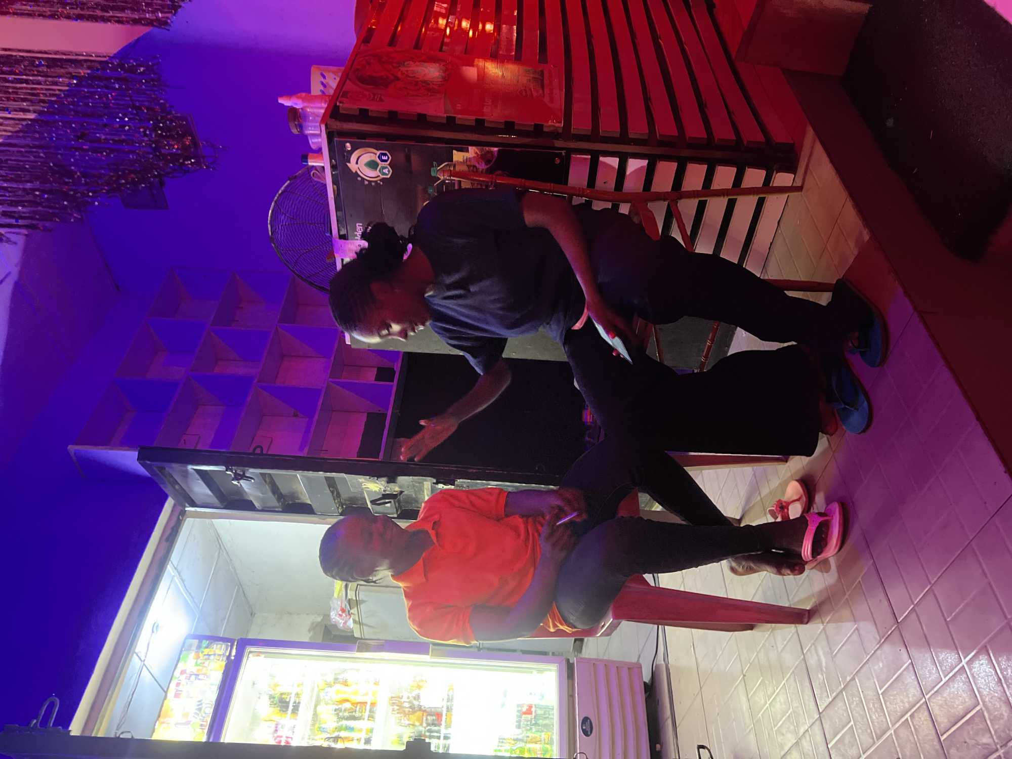 In a dimly lit indoor setting in Howayi, Nigeria, two dark-skinned individuals are seated on stools, engaged in conversation. The scene is illuminated by artificial purple and red lighting, with a bright white light emanating from a commercial refrigerator on the far left.
The individual on the left is seated on a pink plastic stool, wearing an orange short-sleeved polo shirt and dark pants. They hold a pen or thin object in their right hand and are looking towards the other person with a slight smile. Pink flip-flops rest on the tiled floor near their bare feet.
The individual on the right is seated on a dark red wooden stool, wearing a dark blue t-shirt and dark pants, with blue flip-flops on their feet. Their right arm is raised in a gesturing motion, and they hold a phone in their left hand, directing their gaze towards the first individual. Their braided hair is pulled back into a bun.
The background features a white commercial refrigerator displaying bottles of beverages on the far left. An open doorway above the refrigerator reveals a light-colored wall and ceiling. To the right, a dark counter or cabinet is topped with a large industrial fan and various small containers. This structure displays a logo with a green leaf design and the partial word "den." Further to the right, a red slatted partition forms a wall, and above, decorative glittery streamers hang from the ceiling. The upper right wall features a series of cubby-like shelves. The floor is covered in light-colored tiles, reflecting the ambient purple and red lighting.