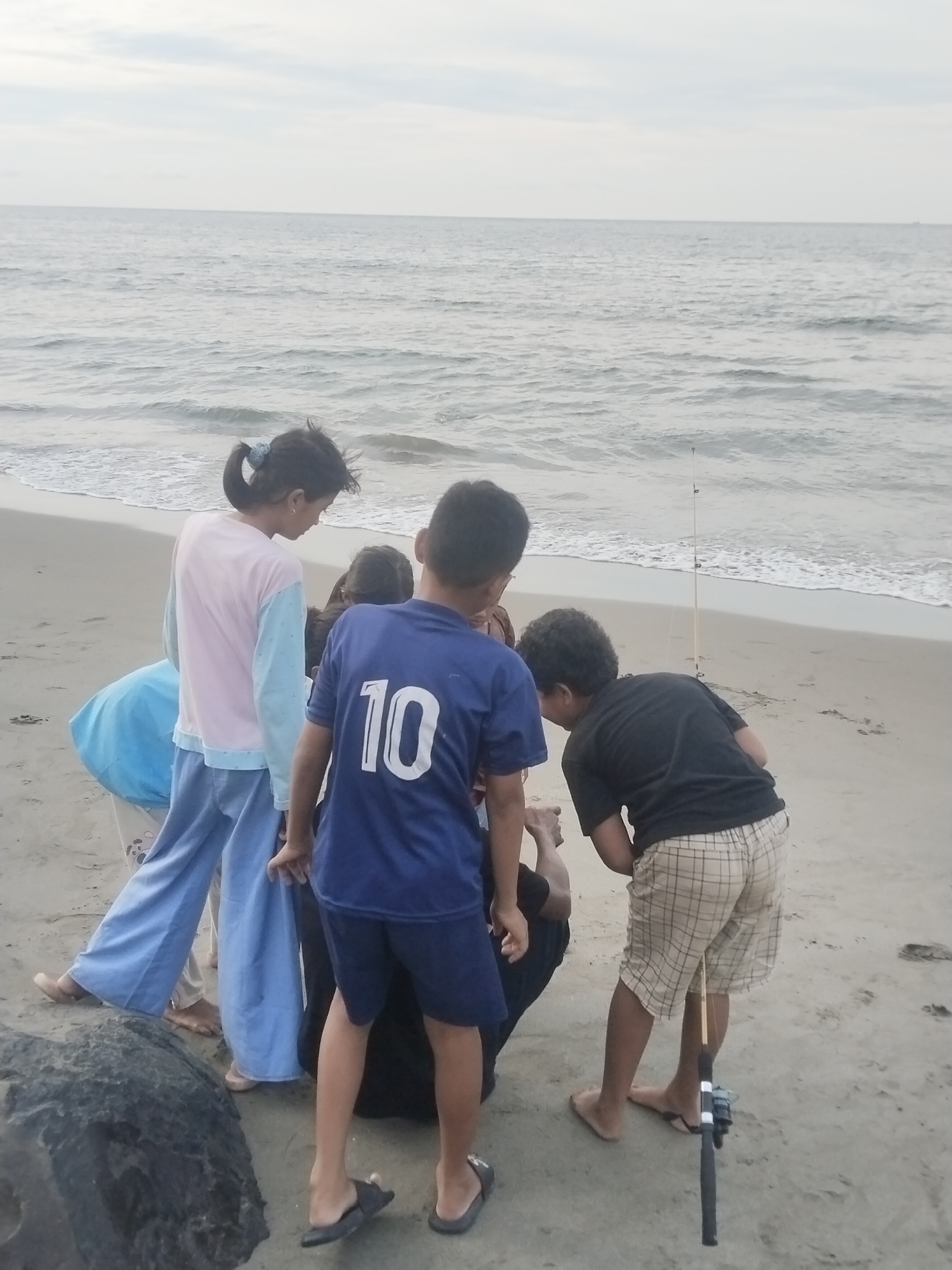 A group of at least five children are gathered on a sandy beach in Lhokseumawe, Indonesia, with the ocean and a pale sky in the background. A fishing rod is propped up in the sand to the right of the frame. In the foreground, a large dark rock is visible on the left. The children are casually dressed and appear to be interacting with each other, some looking down towards the sand and others towards the water. One child in the center wears a blue t-shirt with the number "10" on the back. Another child to the left wears a light pink and blue long-sleeved shirt and loose blue pants.