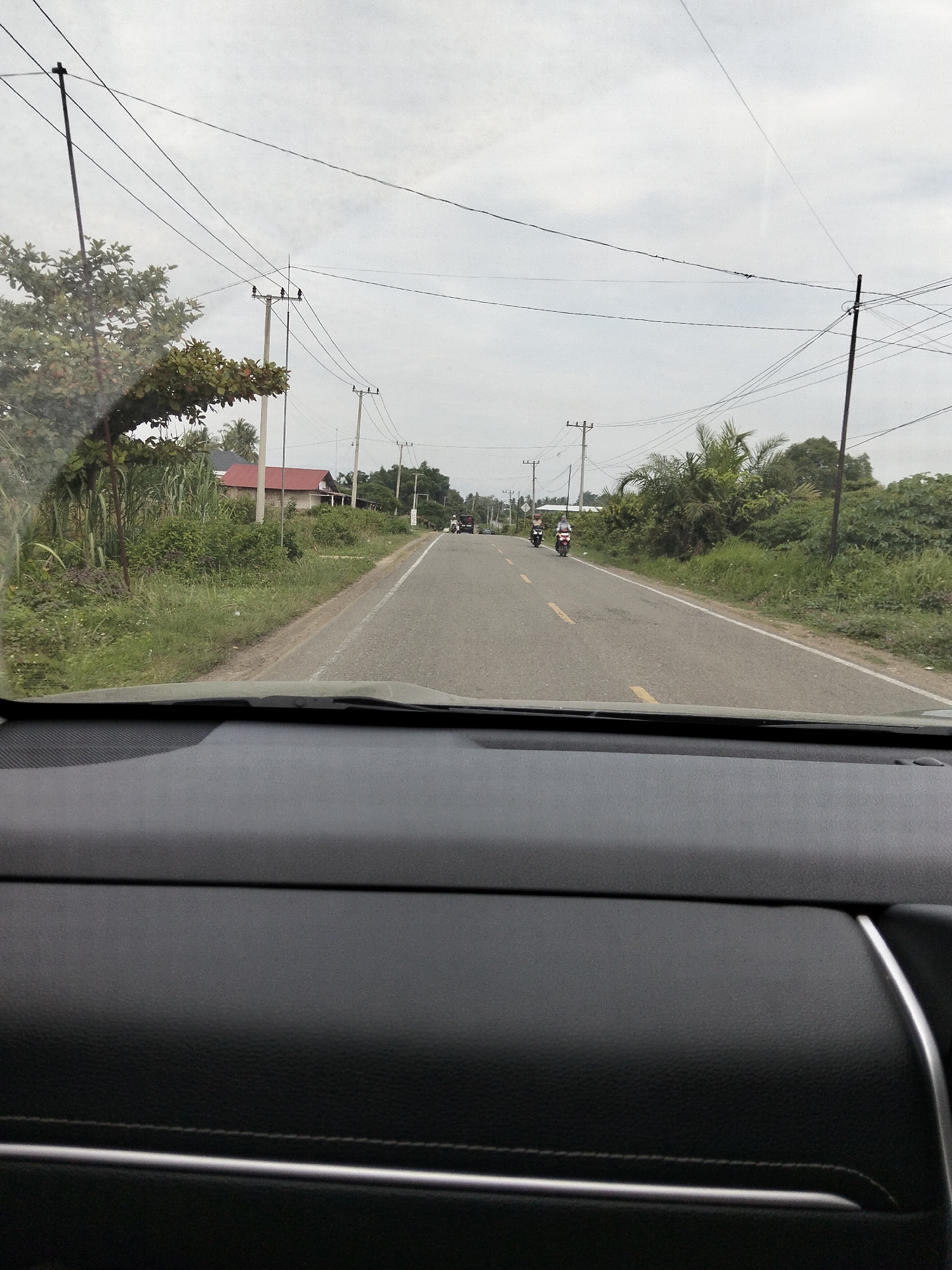 This image, taken from inside a vehicle, captures a view of a two-lane asphalt road in Tanjongkeumala, Indonesia. The road is bordered by lush green vegetation, including trees and tall grasses. Overhead, a complex network of power lines crisscrosses the sky, supported by utility poles. On the left side of the road, a red-roofed building is visible amongst the greenery. Further down the road, two motorcycles with riders are traveling in the same direction. The sky is overcast, suggesting a cloudy day. The foreground of the image shows the interior of the vehicle, with the dashboard and windshield visible.